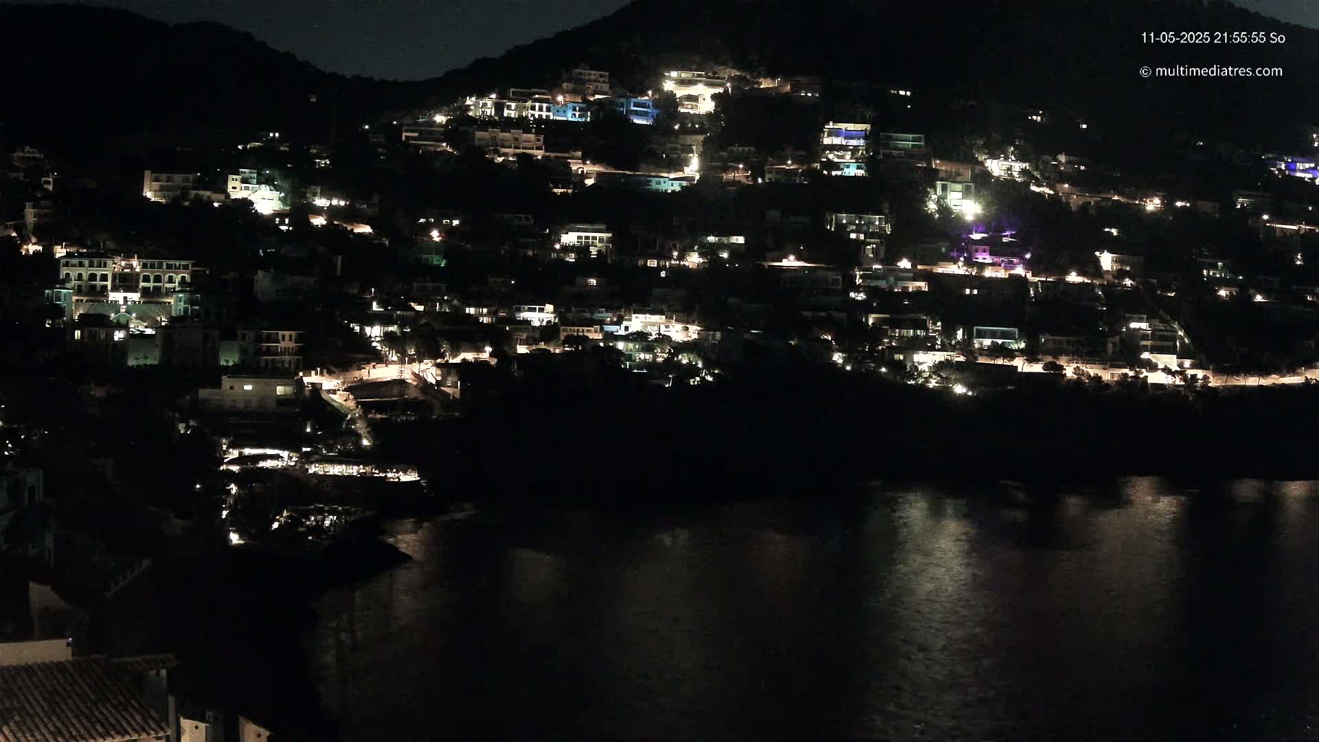 A hillside town is illuminated at night, overlooking a dark body of water.