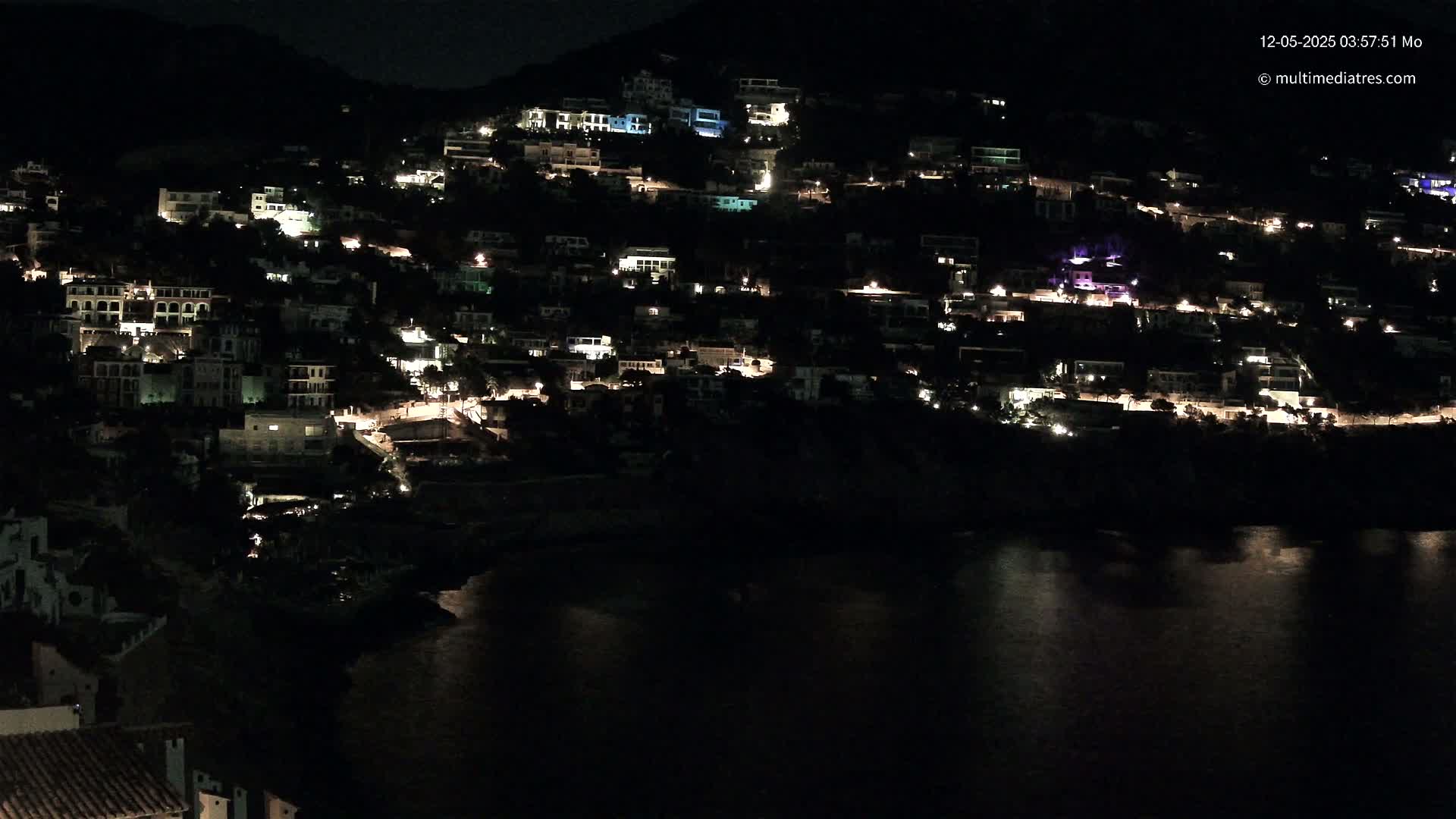 A hillside town at night, with numerous houses illuminated, overlooks a dark body of water.