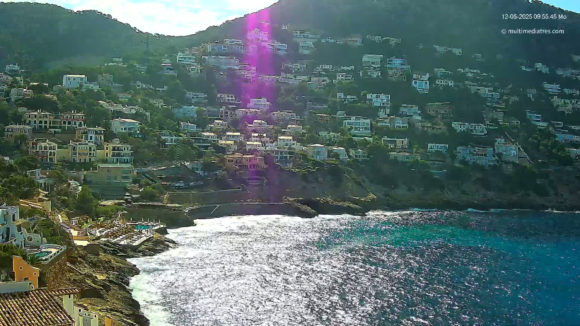 A hillside densely populated with houses slopes down to a rocky coast where waves lap against the shore on a sunny day.