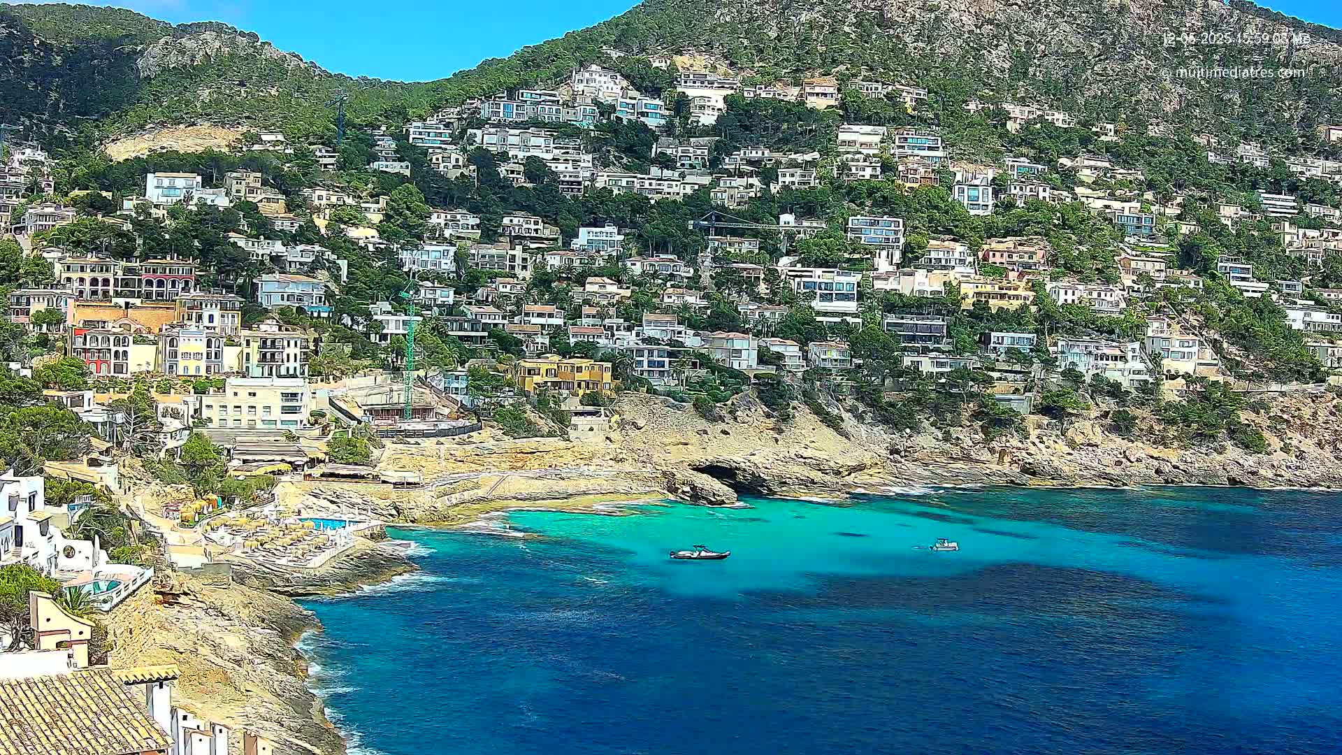 A hillside densely covered with houses slopes down to a clear, turquoise cove with a few small boats.