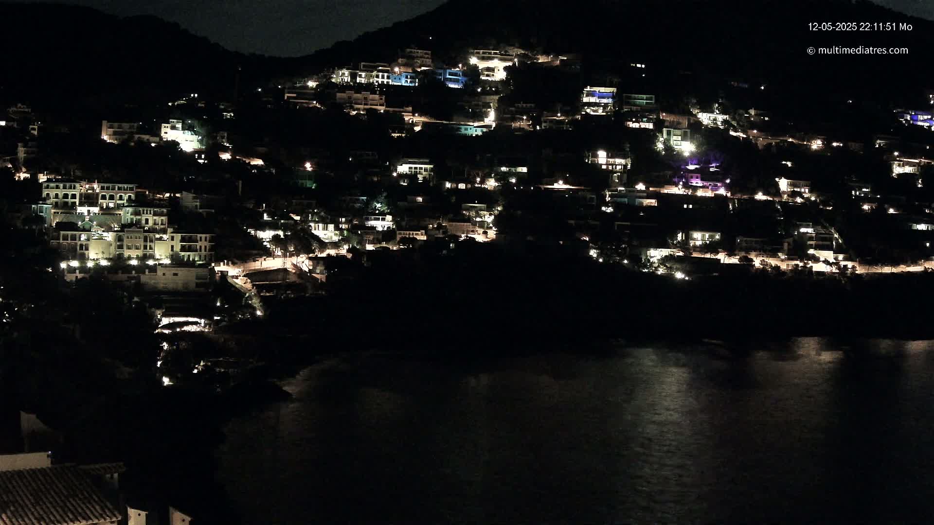 A hillside town at night, illuminated by the lights of numerous houses, overlooks a dark body of water under a clear, night sky.