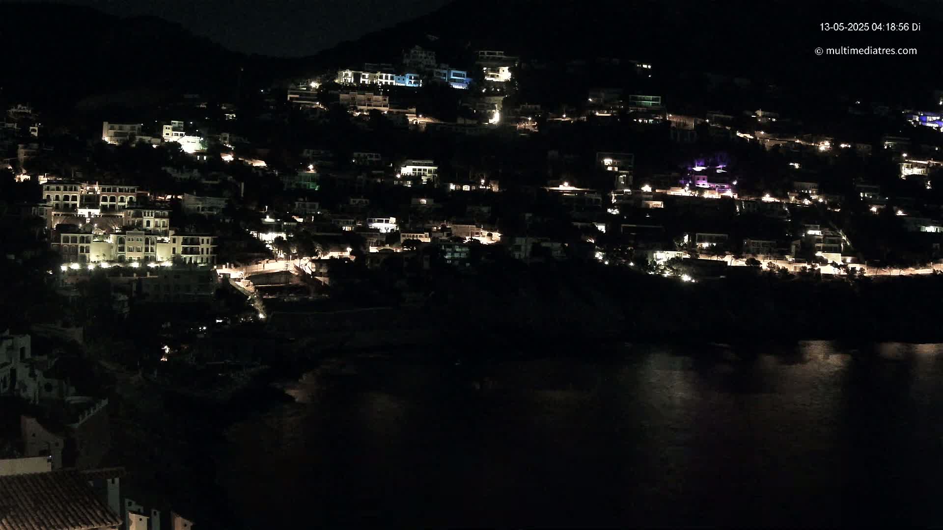 A hillside town at night, illuminated by the lights of many buildings, overlooks a dark body of water.