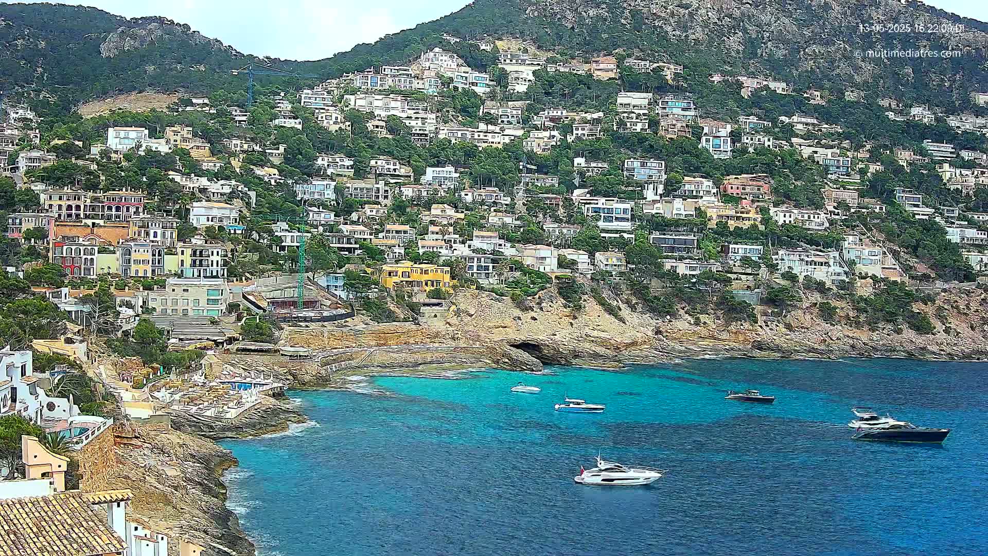 A hillside covered in closely packed houses overlooks a calm, turquoise bay with several boats.