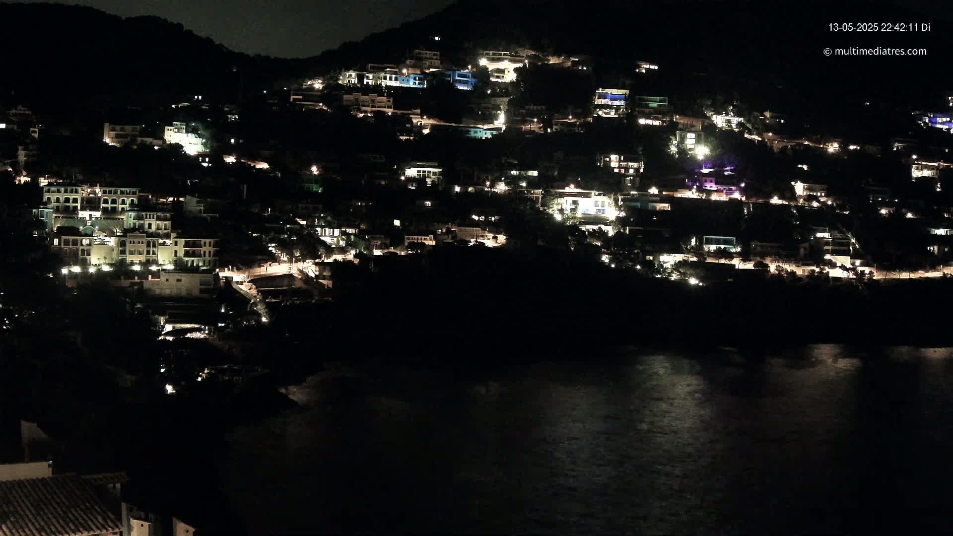 A hillside village at night, illuminated by numerous lights from buildings, overlooks a dark body of water under a clear, dark sky.