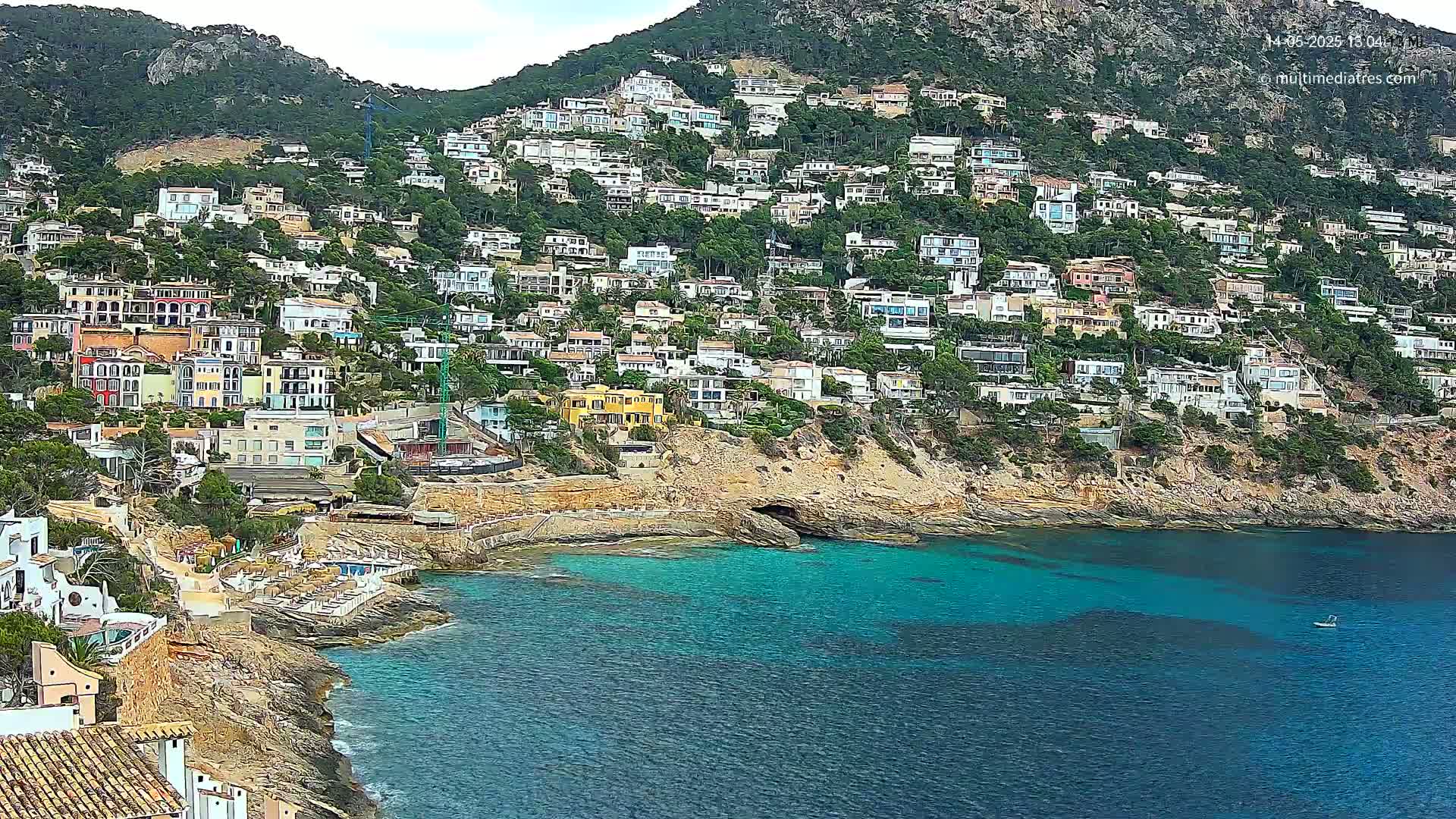 A hillside town of colorful houses on a rocky coast with a calm, teal-colored sea under partly cloudy skies.