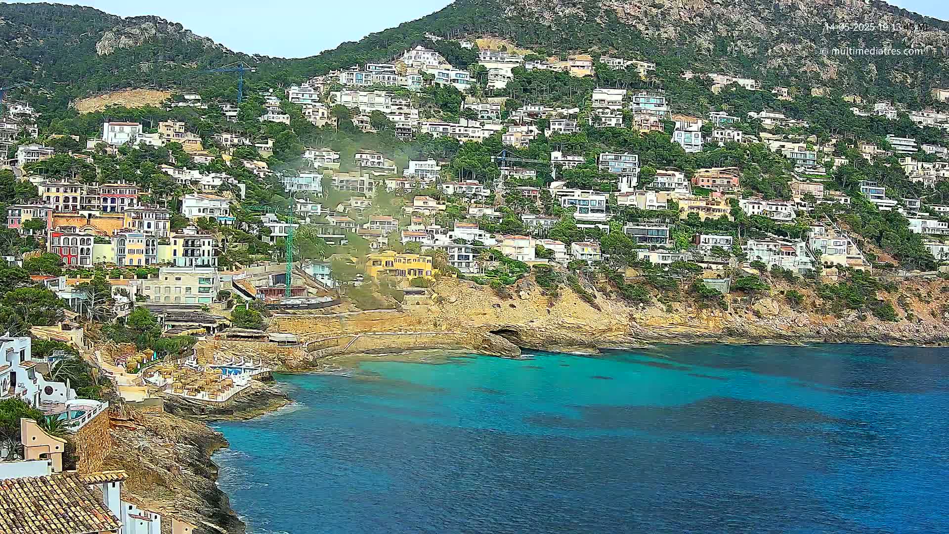A hillside densely packed with houses overlooks a calm, teal-colored cove on a partly cloudy day.