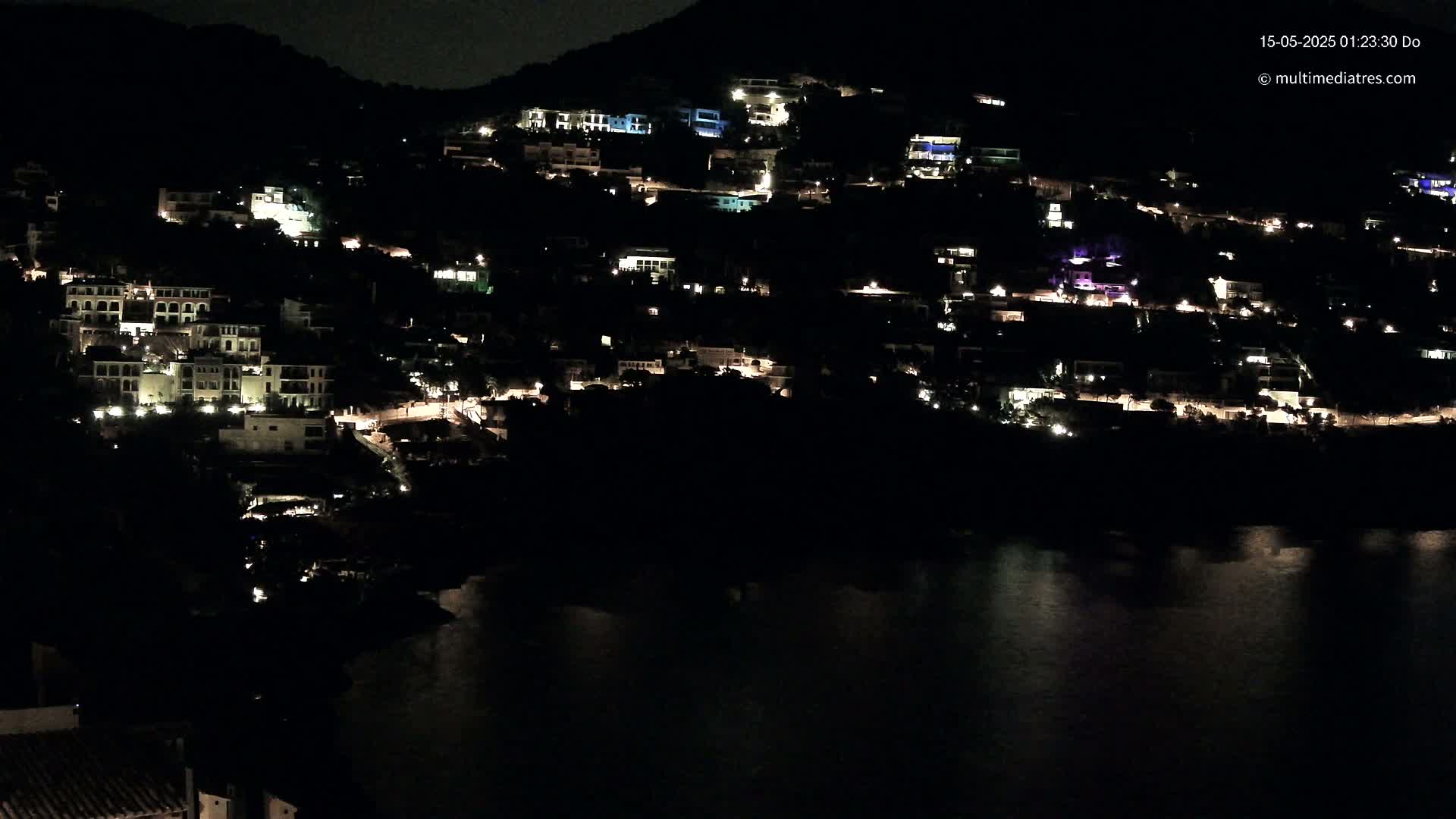 A hillside town at night, with many houses illuminated, overlooks a dark body of water.