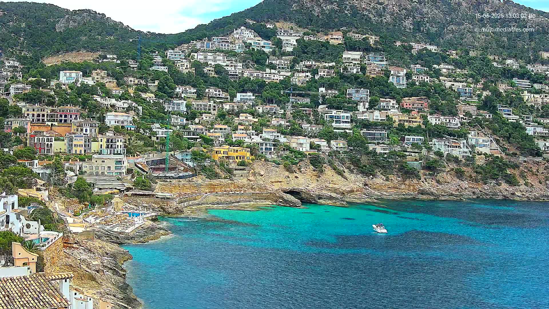 A hillside town of white and colorful houses clings to a rocky coastline, with a small boat on the bright teal water below.