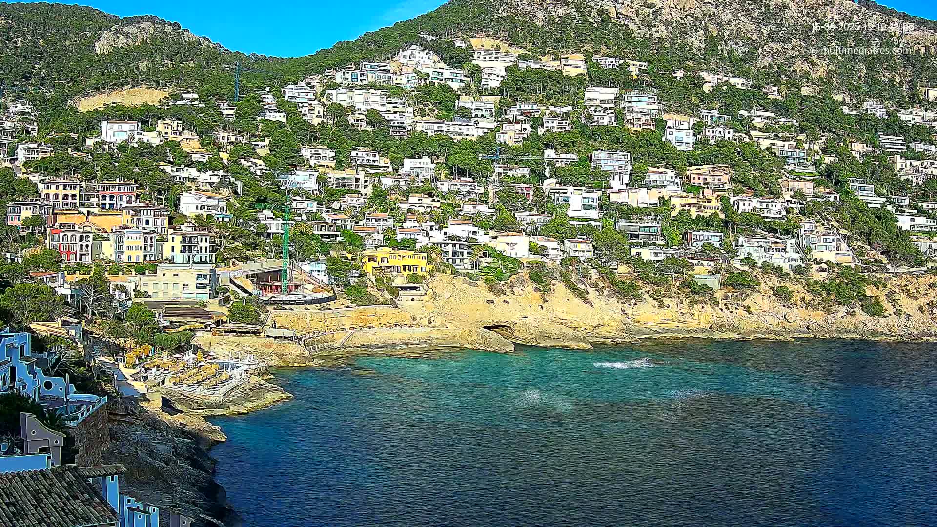 A hillside densely covered with houses descends to a calm, blue-green cove on a sunny day.