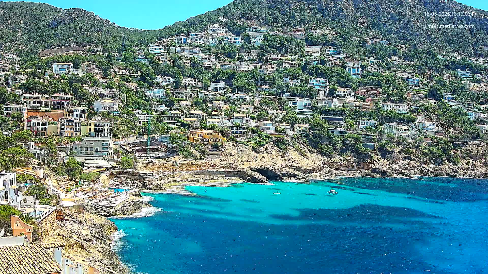 A hillside densely packed with houses overlooks a calm, bright blue ocean cove on a sunny day.