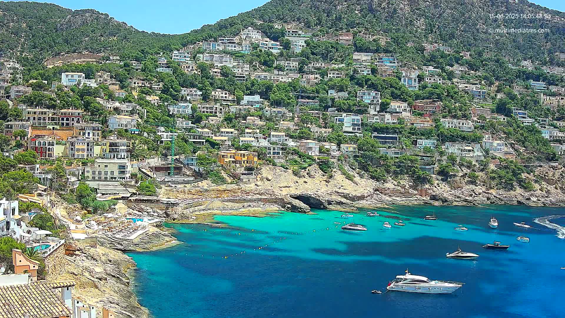 A hillside densely covered with houses overlooks a calm, turquoise bay with several boats at anchor on a sunny day.