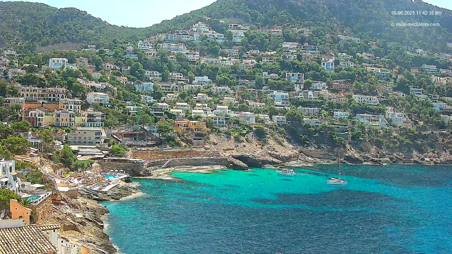 A hillside covered in houses overlooks a calm, turquoise bay with a few boats.