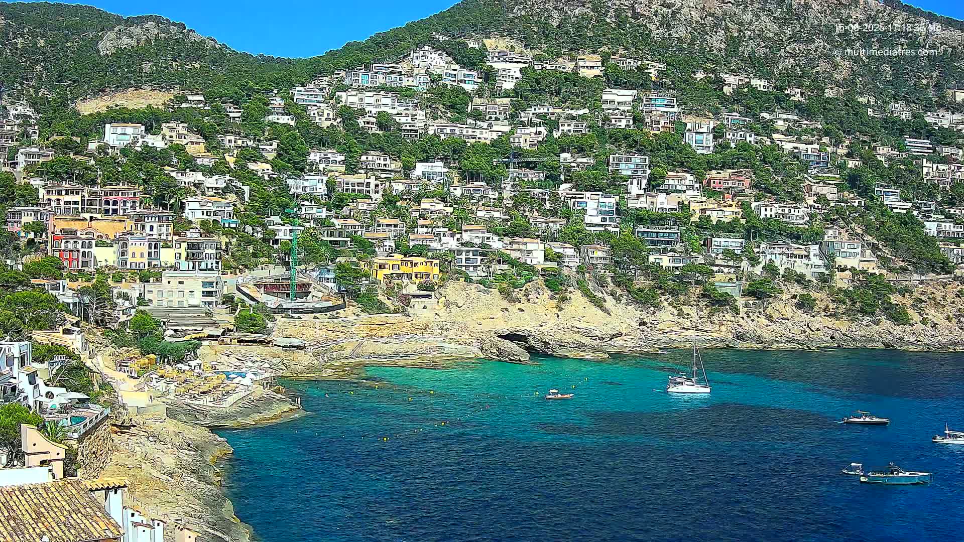 A hillside densely covered with houses overlooks a calm, clear turquoise bay with several boats.