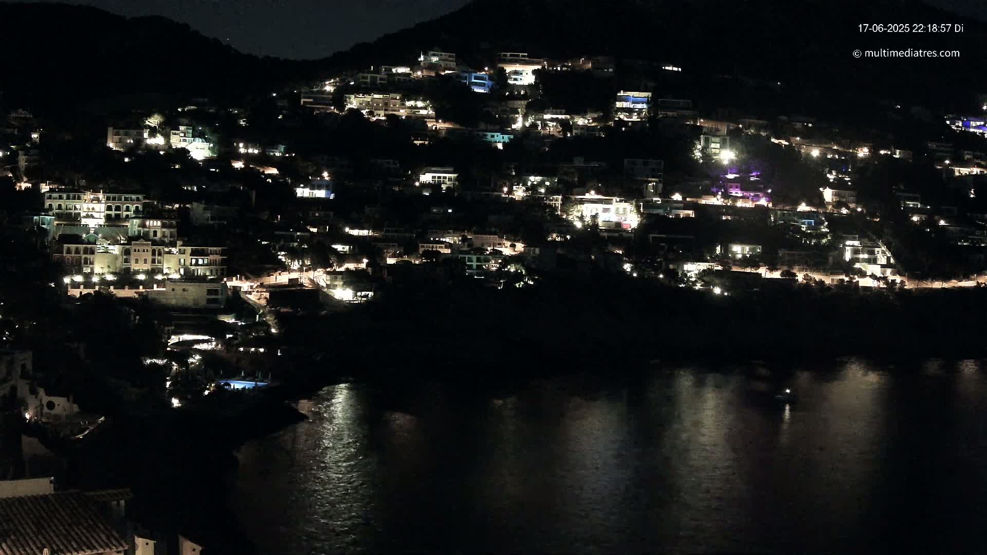 A hillside town at night, illuminated by the lights of numerous buildings, overlooks a dark body of water.