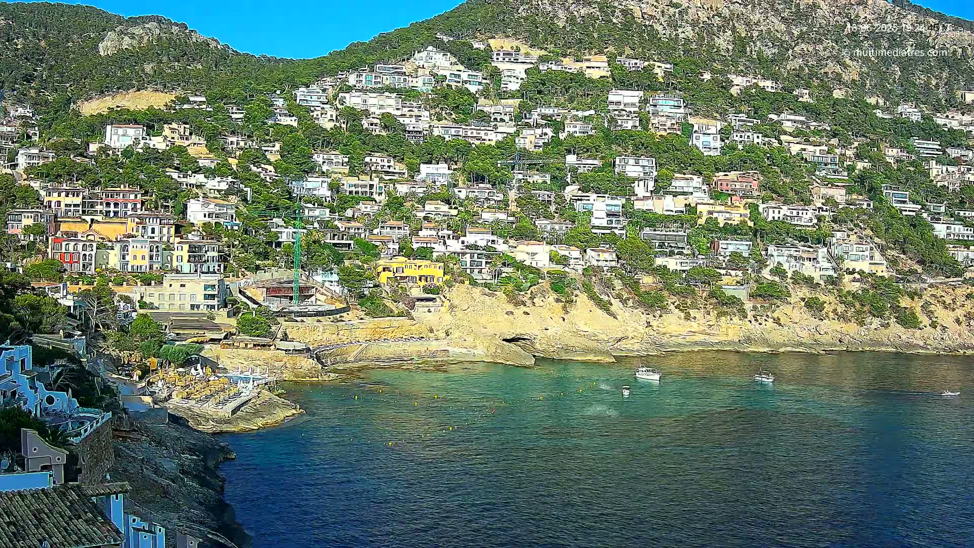 A hillside densely covered with houses slopes down to a calm, blue bay where a few small boats are anchored under a clear, sunny sky.