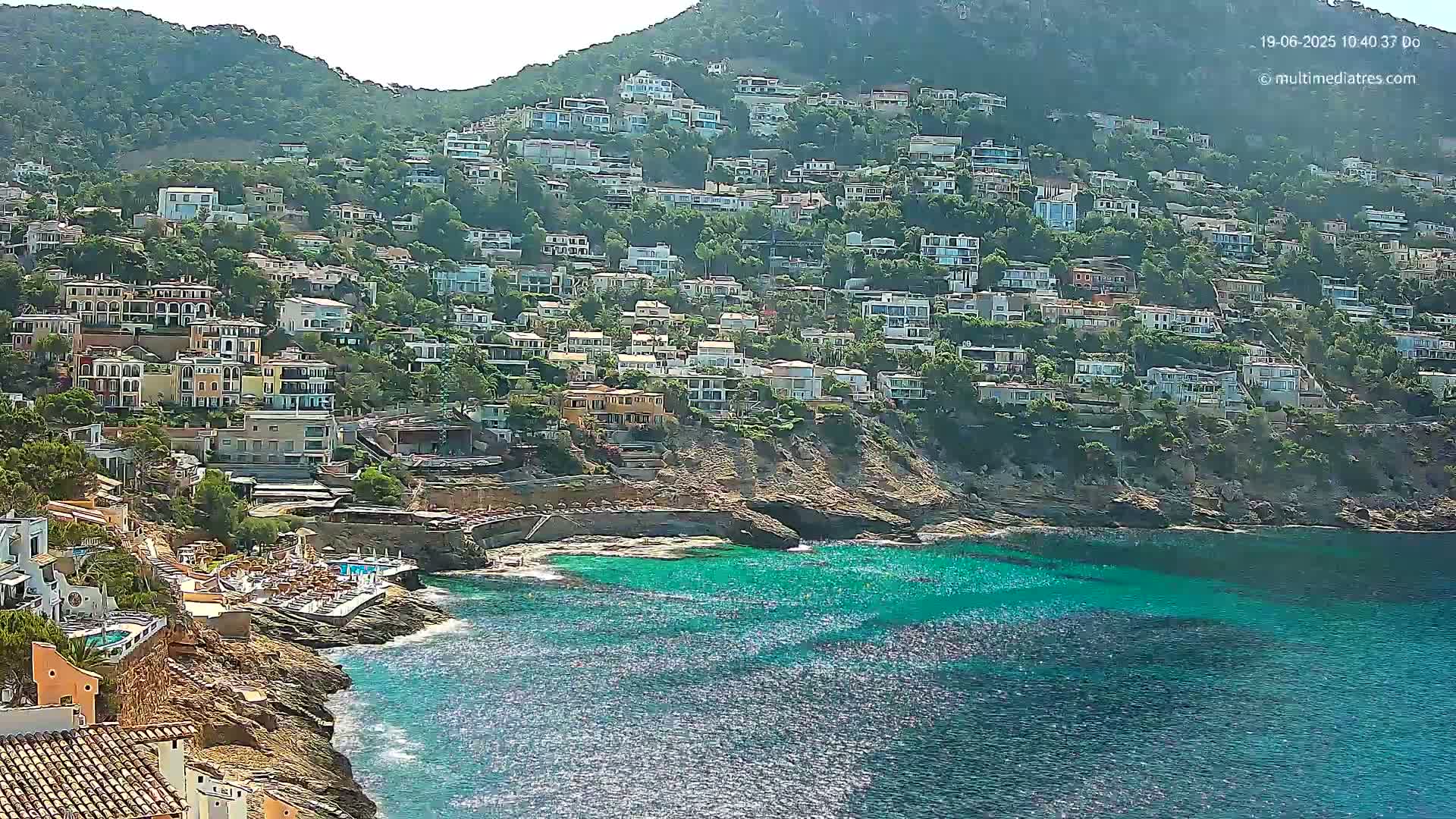 A hillside densely packed with houses overlooks a sparkling turquoise ocean on a sunny day.