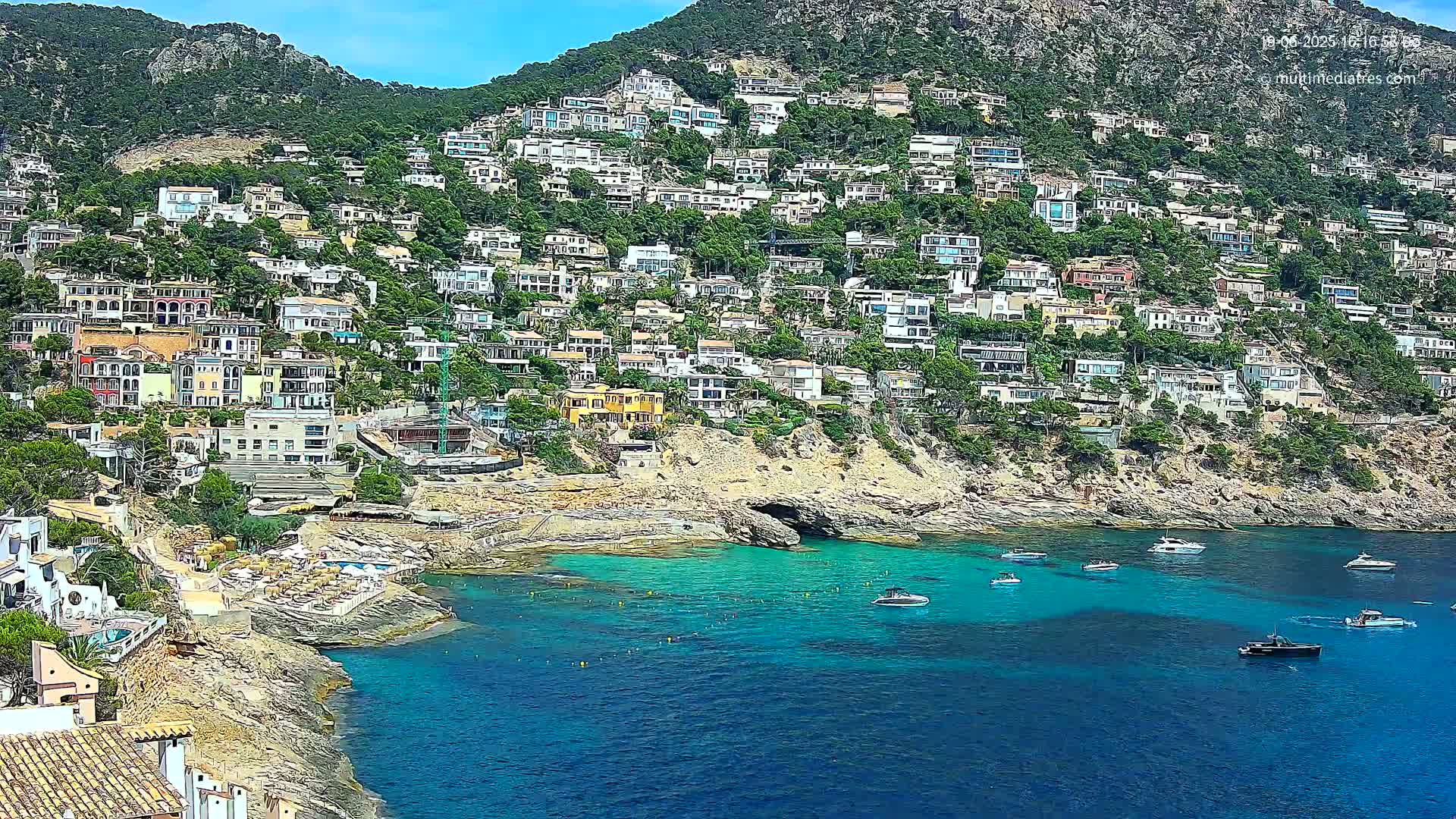 A hillside town with numerous houses overlooks a calm, turquoise bay dotted with boats on a sunny day.