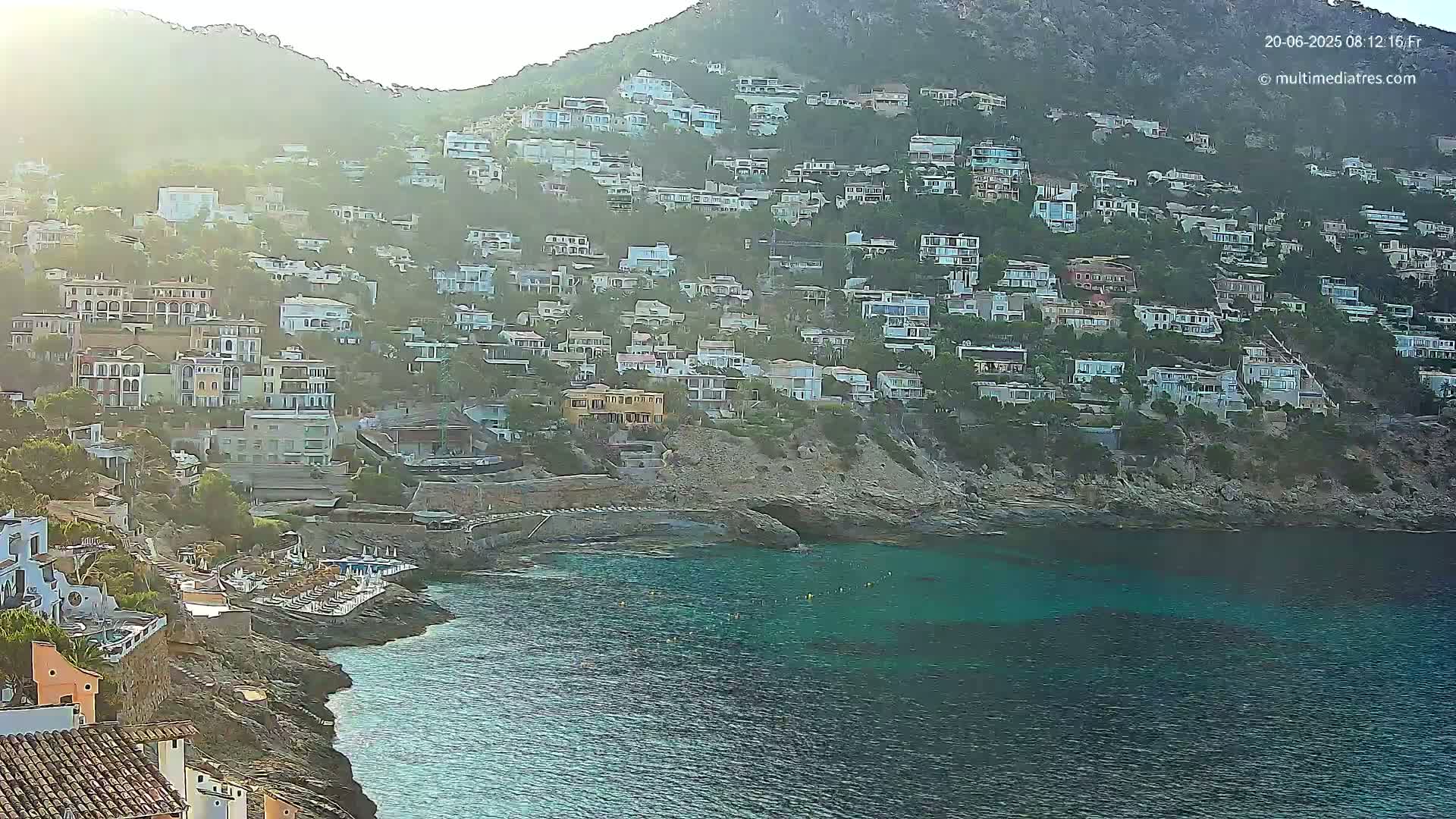 A hillside town of numerous houses sits above a calm, teal cove on a sunny day.