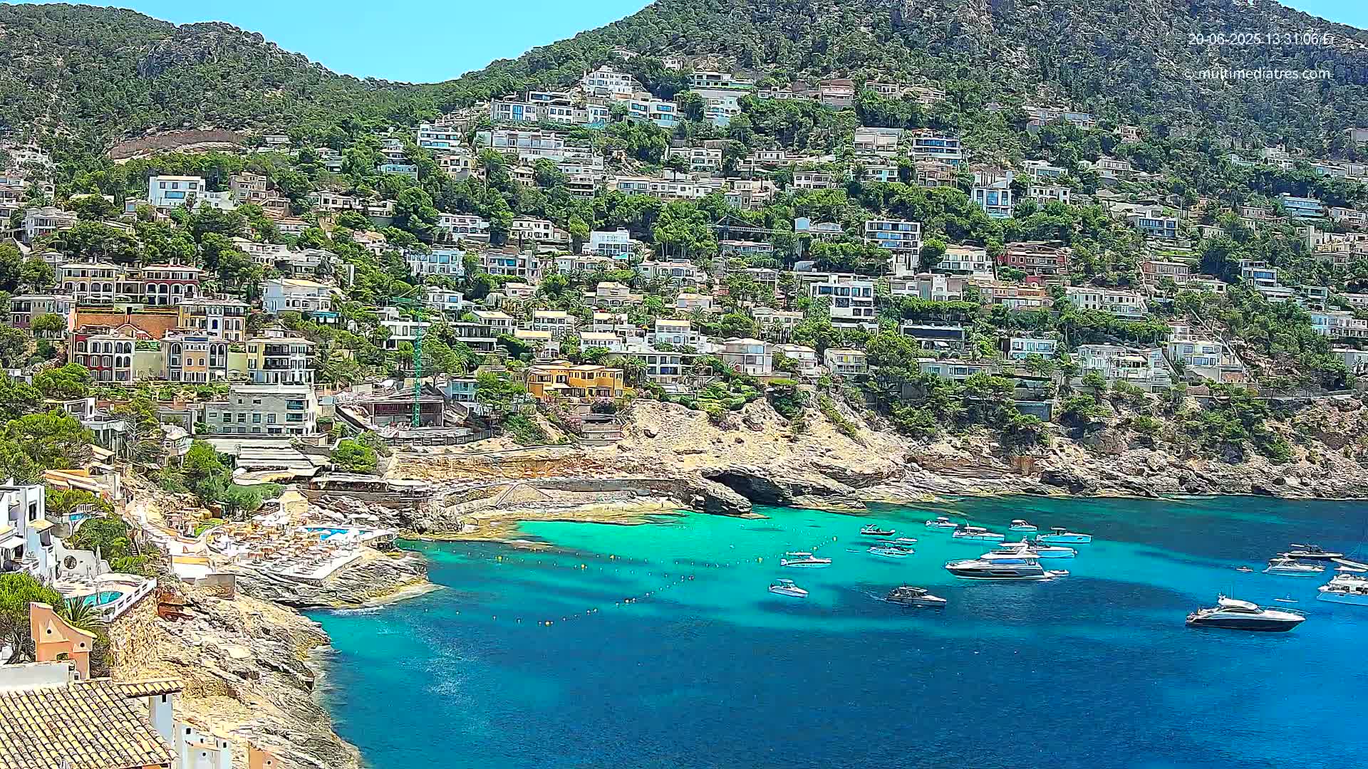 A hillside town of multi-colored houses overlooks a calm, bright blue cove filled with boats on a sunny day.