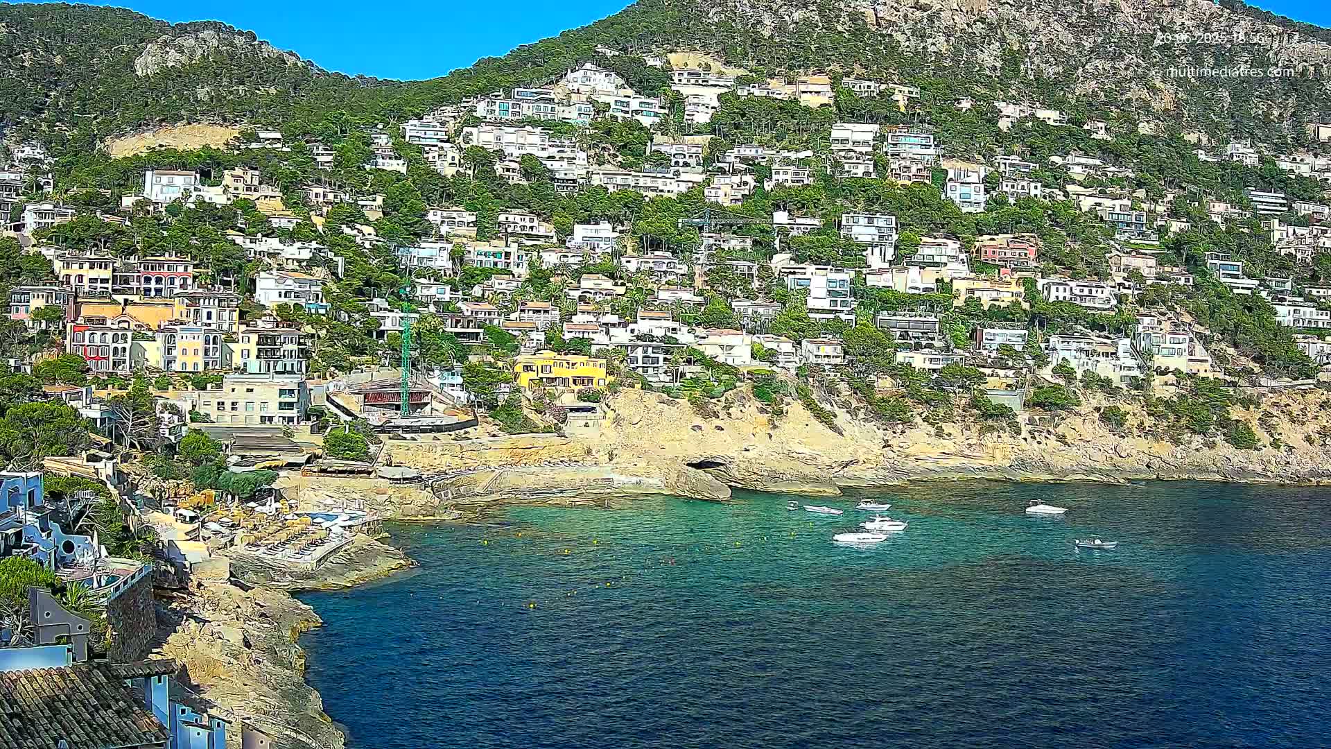 A hillside densely covered with houses overlooks a calm, clear blue bay with several small boats.