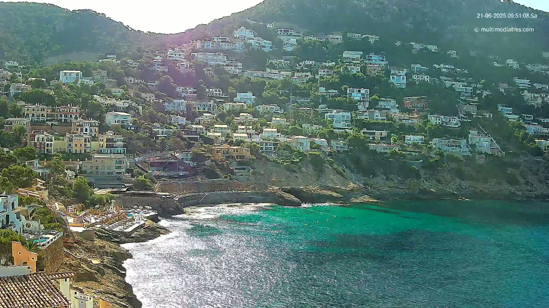 A hillside covered in houses overlooks a calm, turquoise ocean on a sunny day.