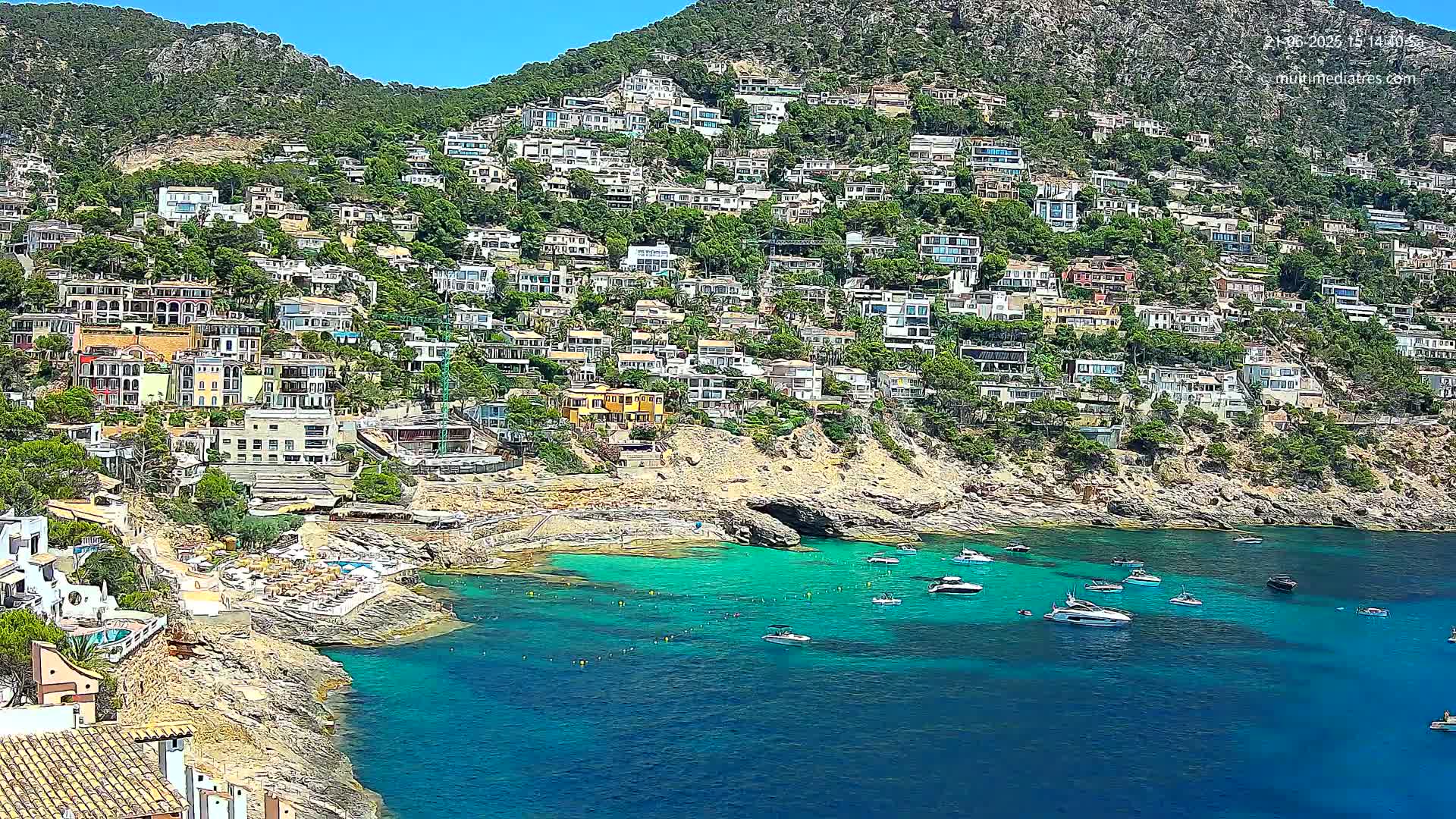 A hillside covered in houses and lush greenery slopes down to a clear, turquoise cove filled with boats on a sunny day.
