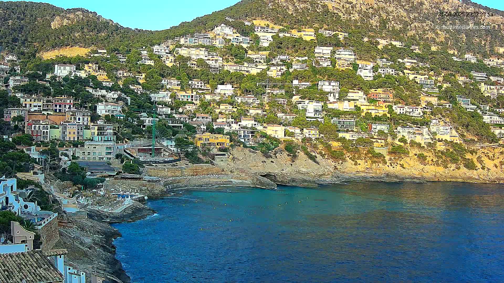 A hillside town of multi-colored houses clings to a rocky coast, overlooking a calm, blue sea under a clear sky.