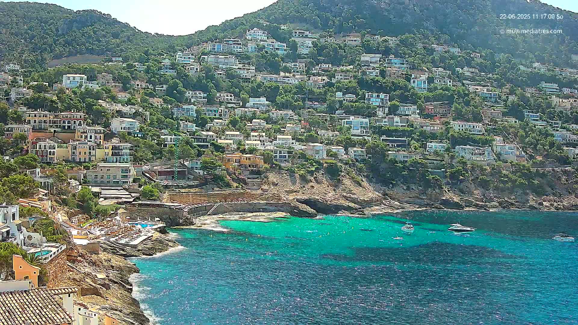 A hillside densely covered with houses overlooks a calm, turquoise bay with several boats anchored near a rocky shore.