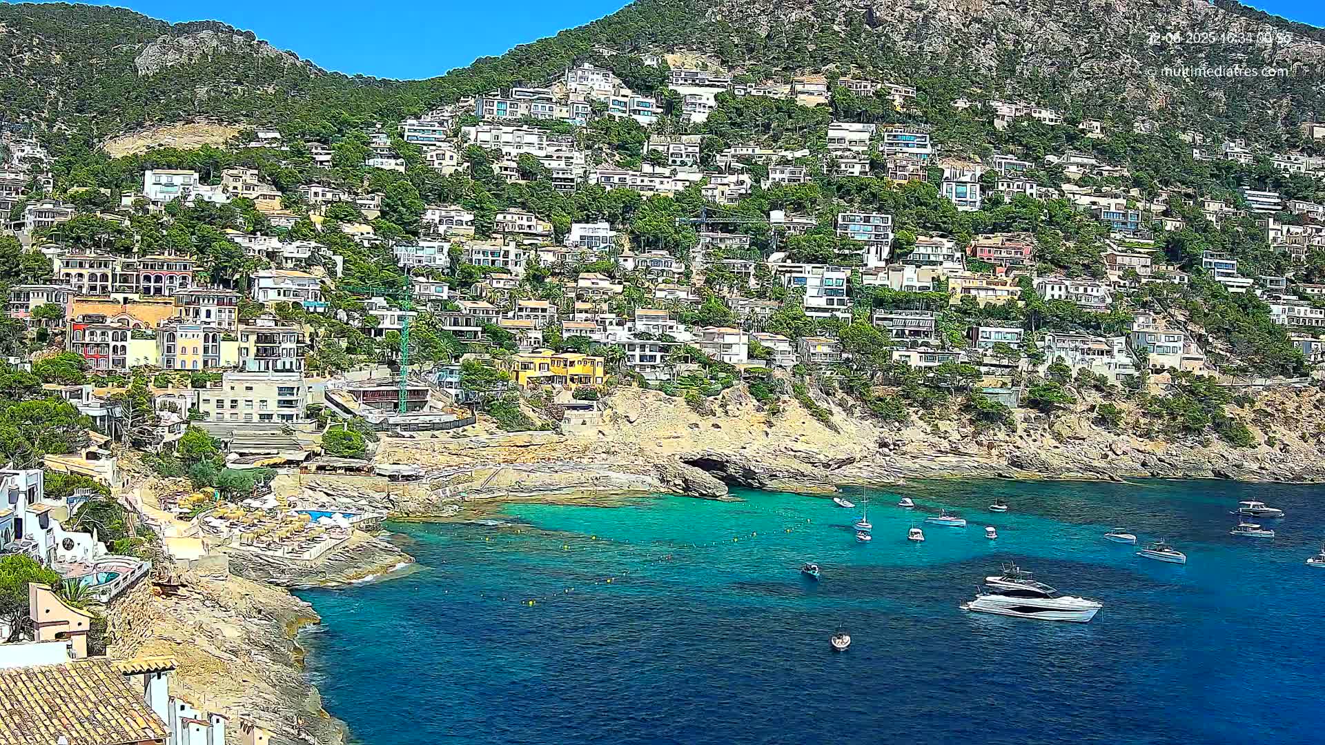 A hillside covered in houses overlooks a calm, turquoise bay with several boats anchored in the clear water under a bright, sunny sky.