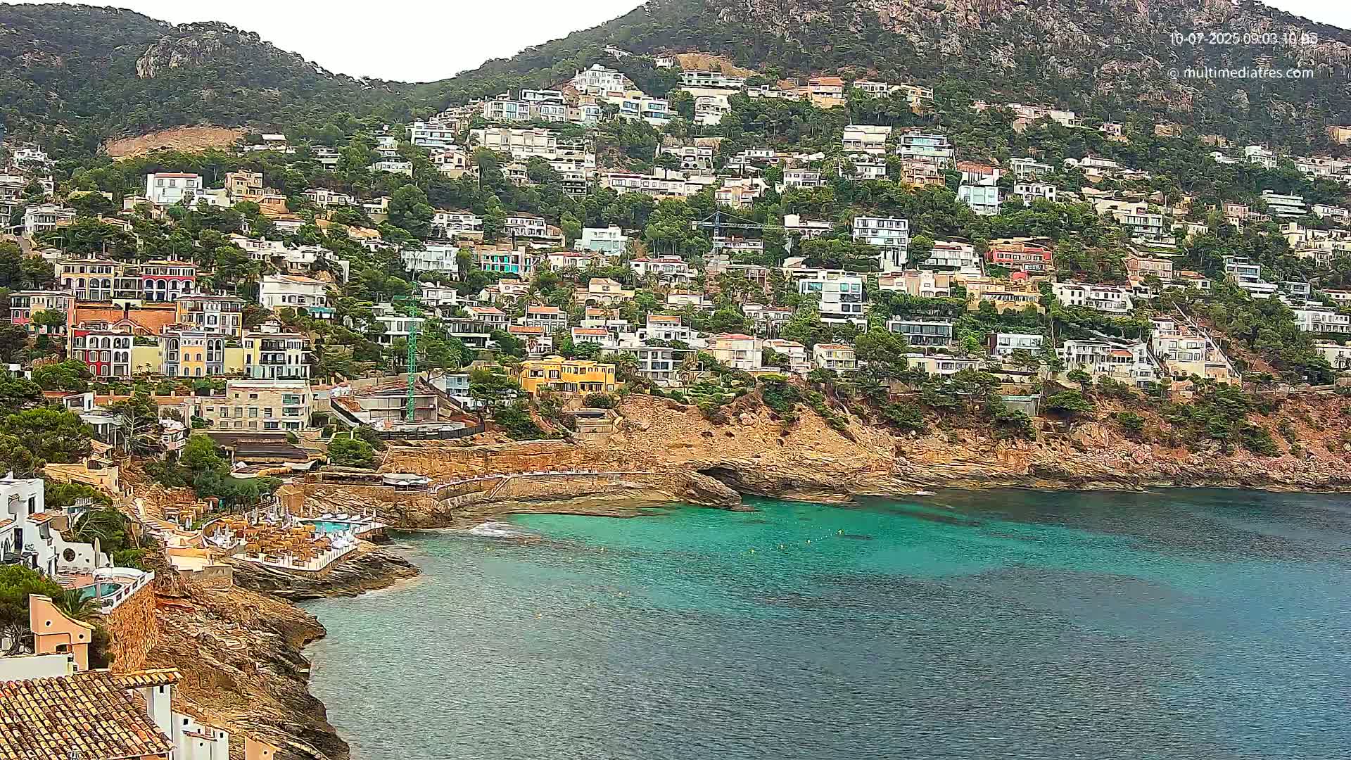 A hillside densely covered with houses overlooks a calm, teal-colored bay on an overcast day.