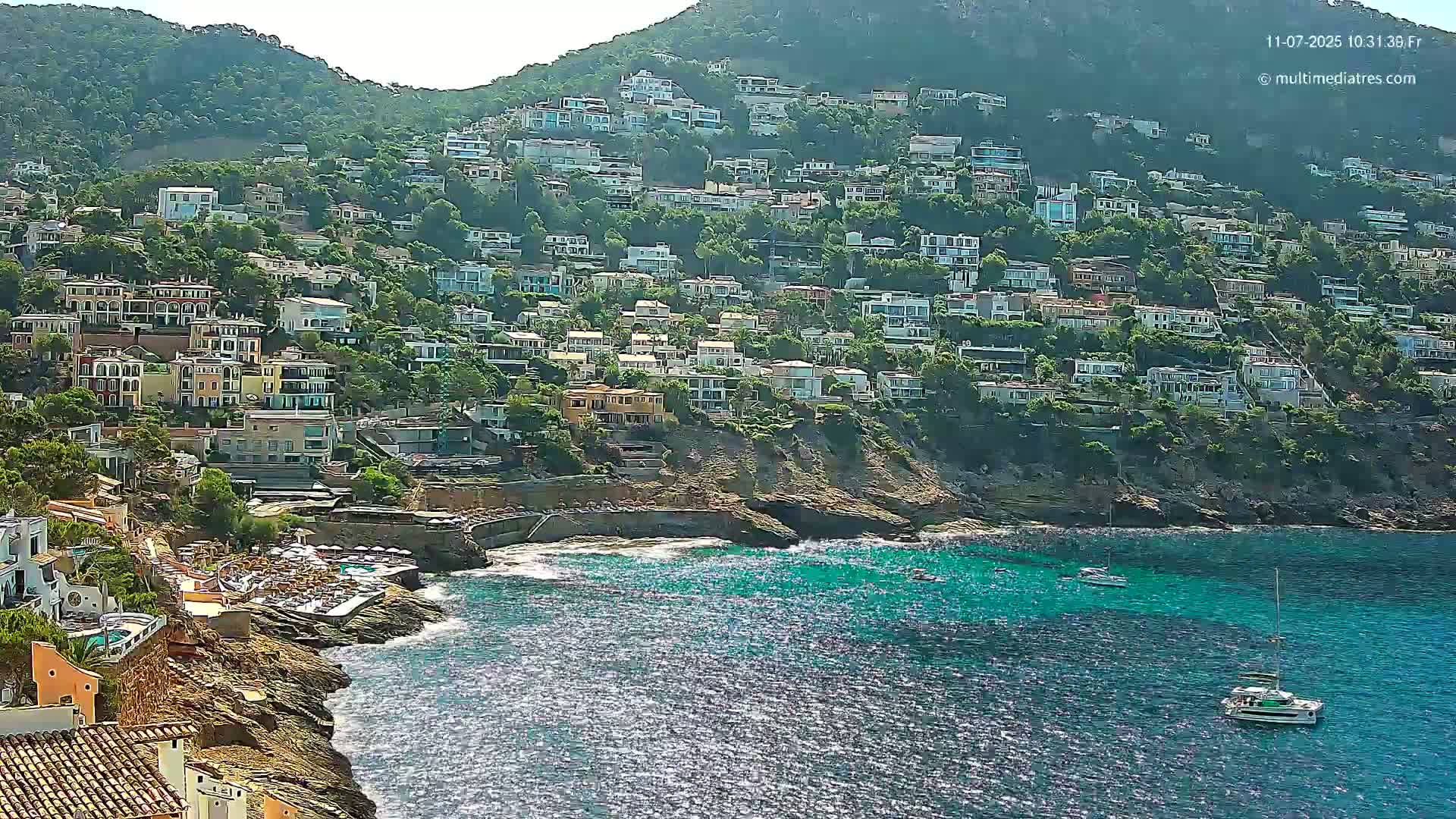 A hillside covered in houses slopes down to a rocky coast with turquoise water, under sunny conditions, and two sailboats are visible in the ocean.