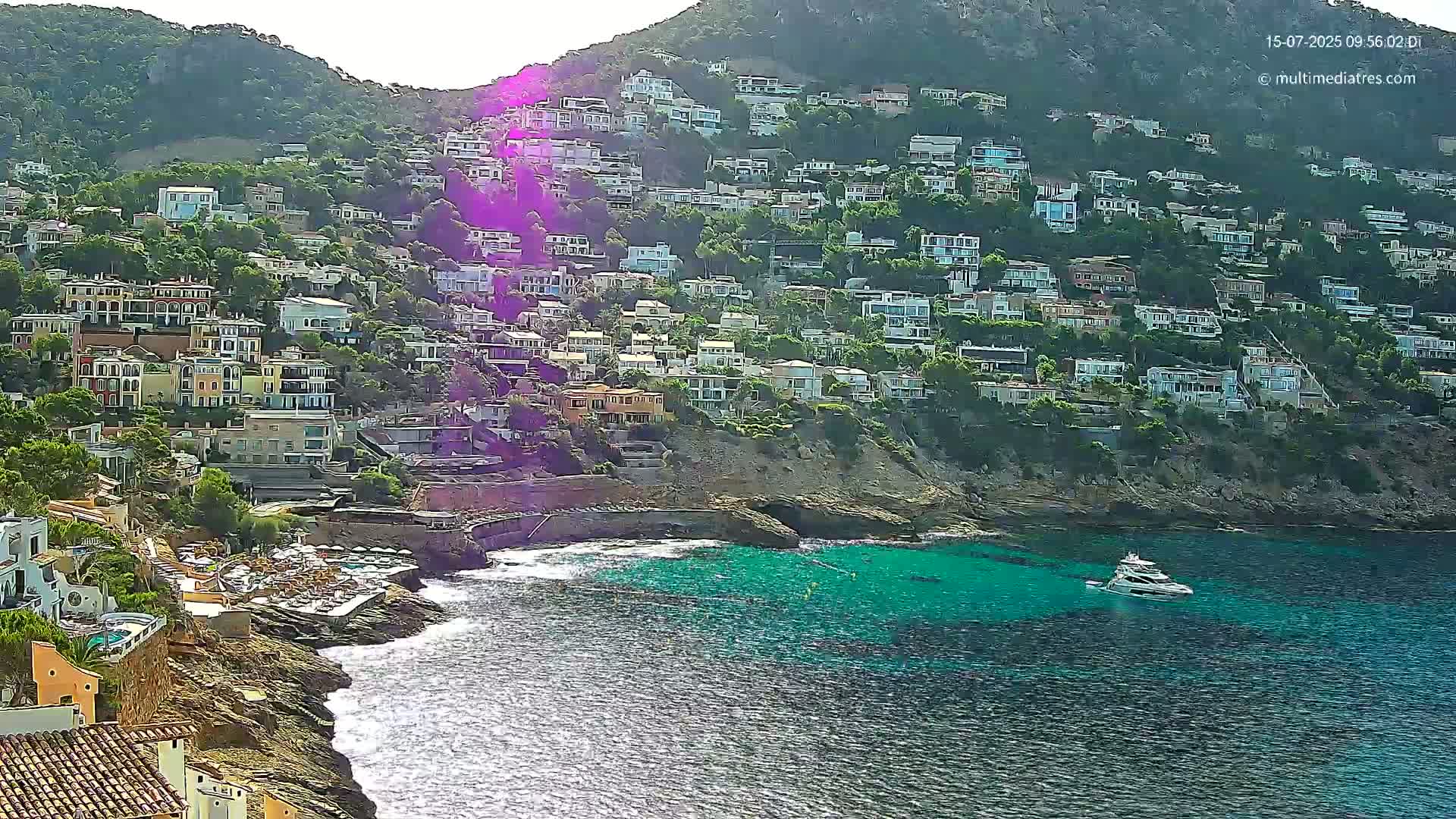 A hillside densely covered with houses slopes down to a rocky coast with turquoise water, where a boat is anchored on a sunny day.