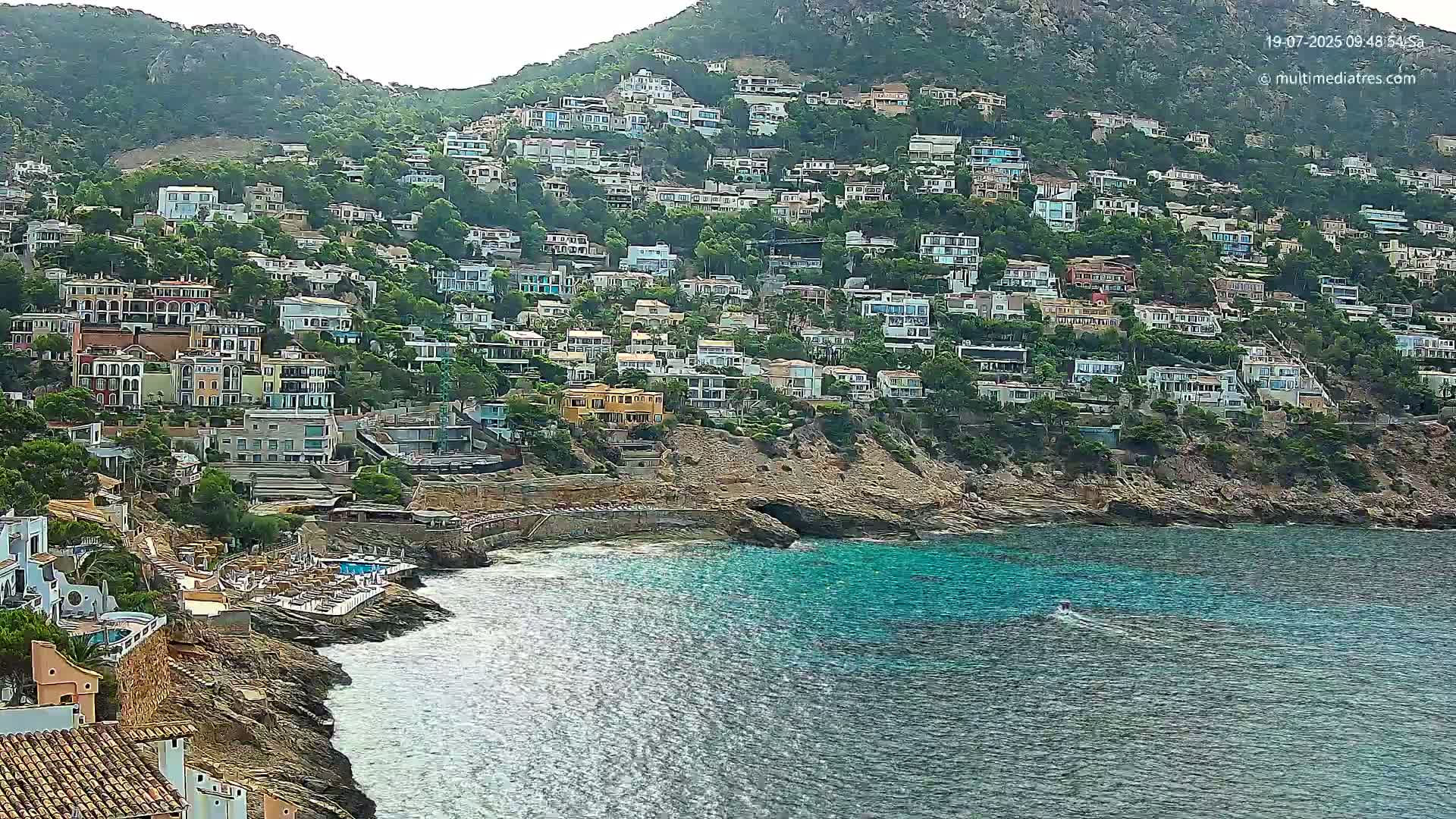 A hillside densely covered with houses slopes down to a rocky coastline with a small, calm bay of turquoise water.