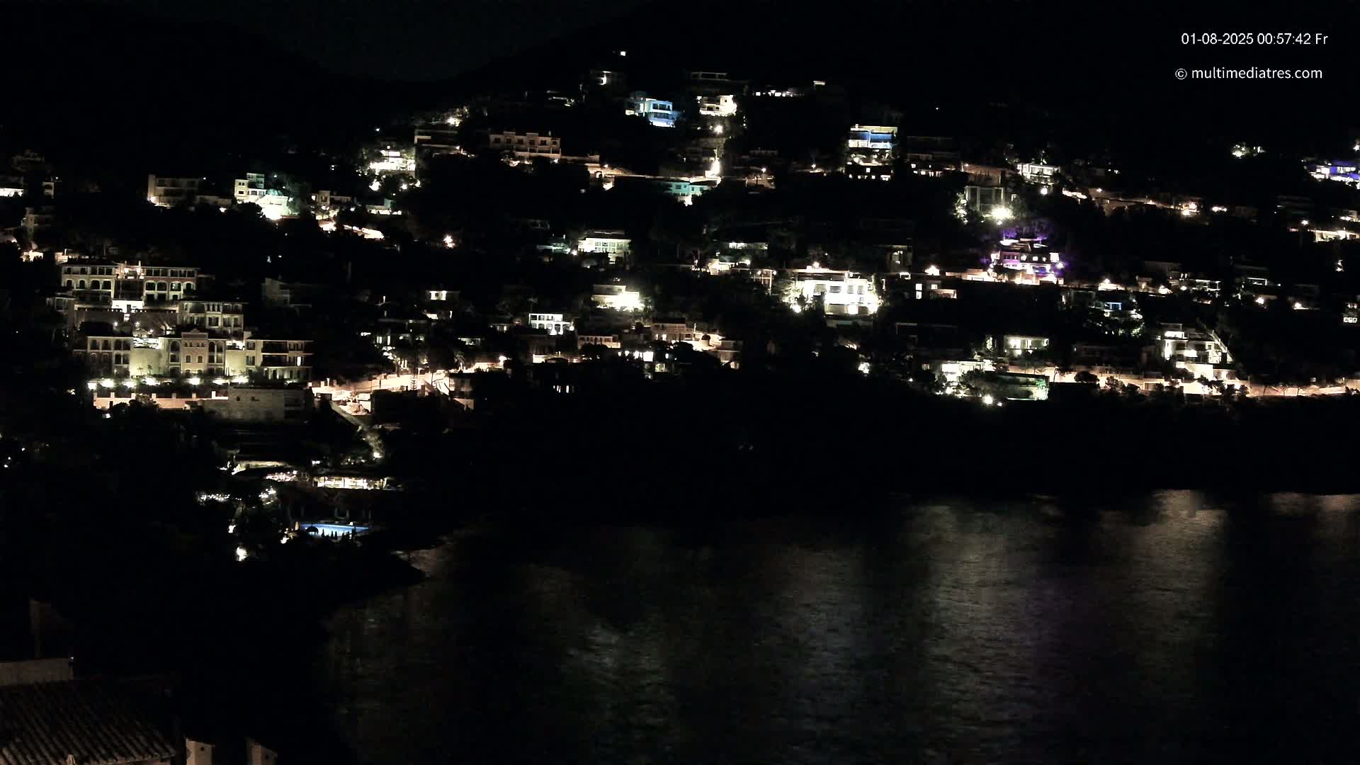 A hillside town at night, illuminated by many house lights, sits above a dark body of water reflecting the lights.