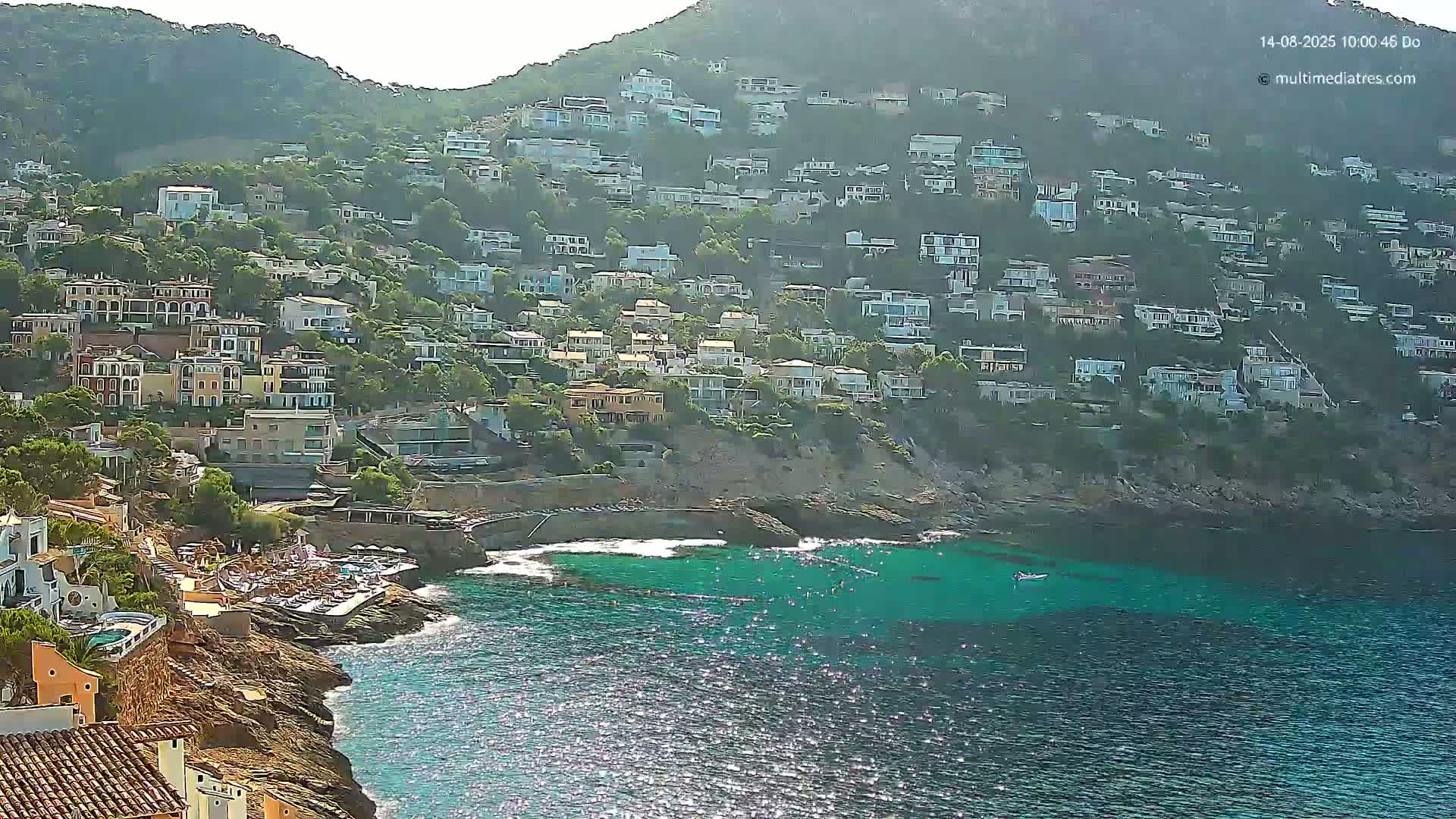 A hillside densely populated with houses overlooks a calm, clear turquoise cove on a sunny day.