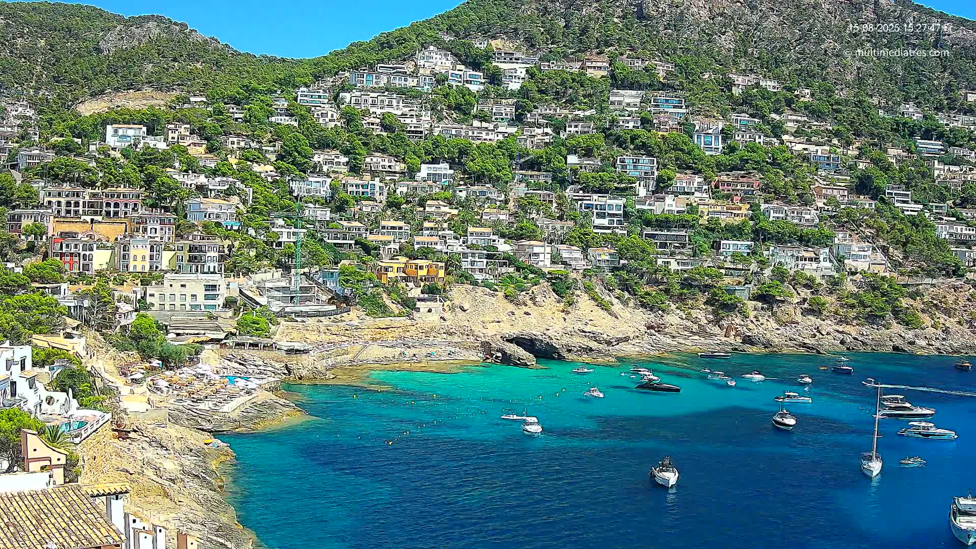 A hillside densely populated with houses overlooks a calm, clear turquoise cove on a sunny day.