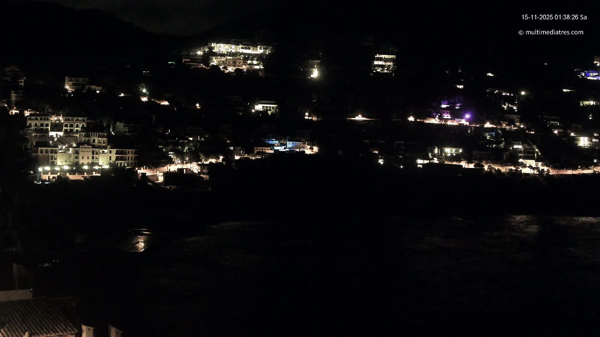 A nighttime panoramic view shows numerous illuminated buildings and scattered streetlights dotting a dark hillside, overlooking a body of water with faint reflections, under clear skies.
