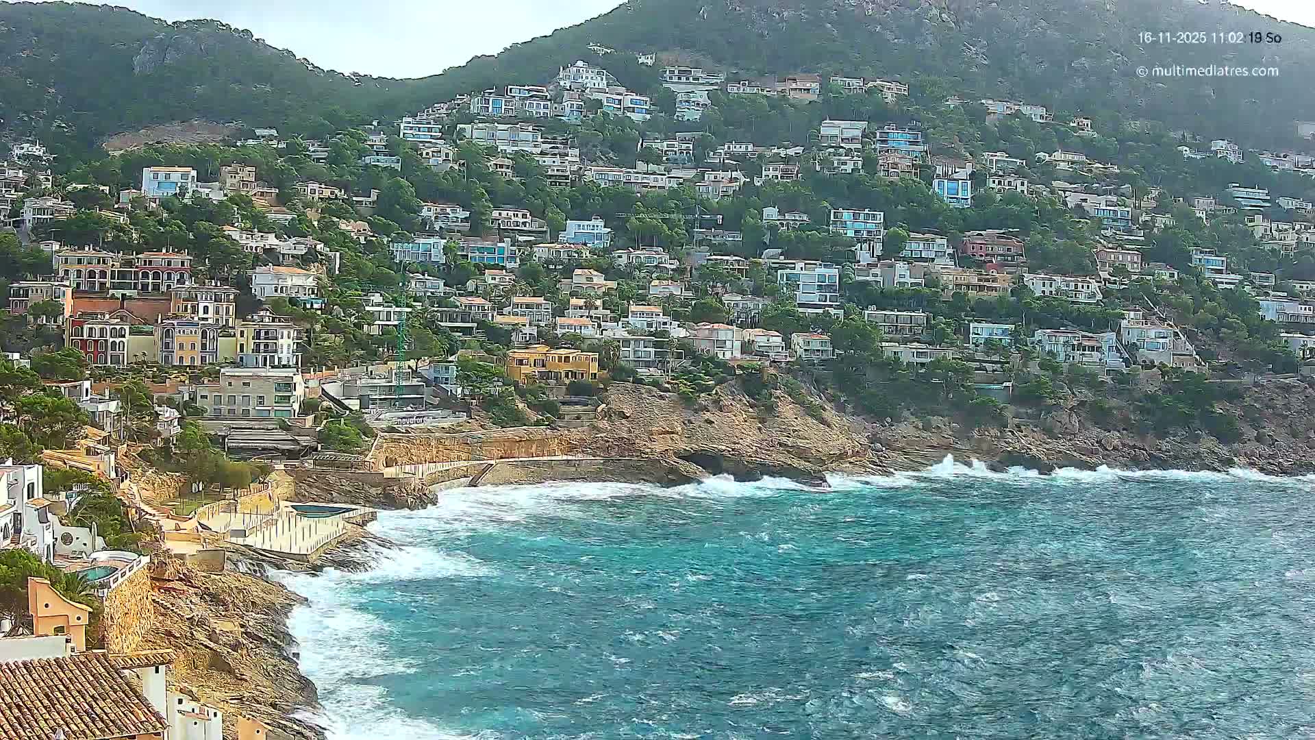 A steep, green hillside densely packed with numerous multi-colored houses descends to a rugged rocky shoreline, where choppy turquoise waves crash against the rocks under a bright, overcast sky.