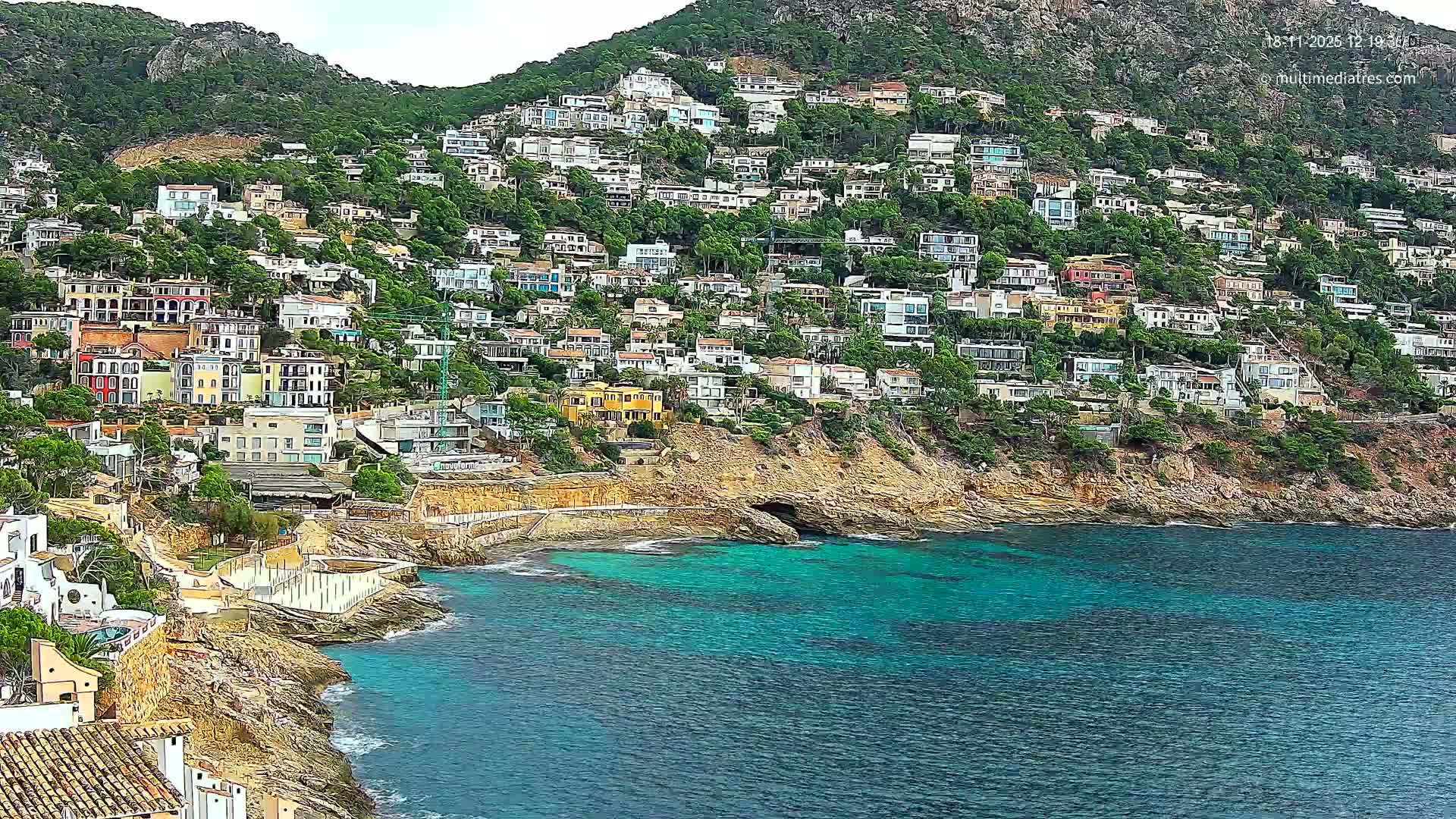 A steep, green hillside densely packed with numerous multi-colored houses descends to a rugged rocky shoreline, where choppy turquoise waves crash against the rocks under a bright, overcast sky.