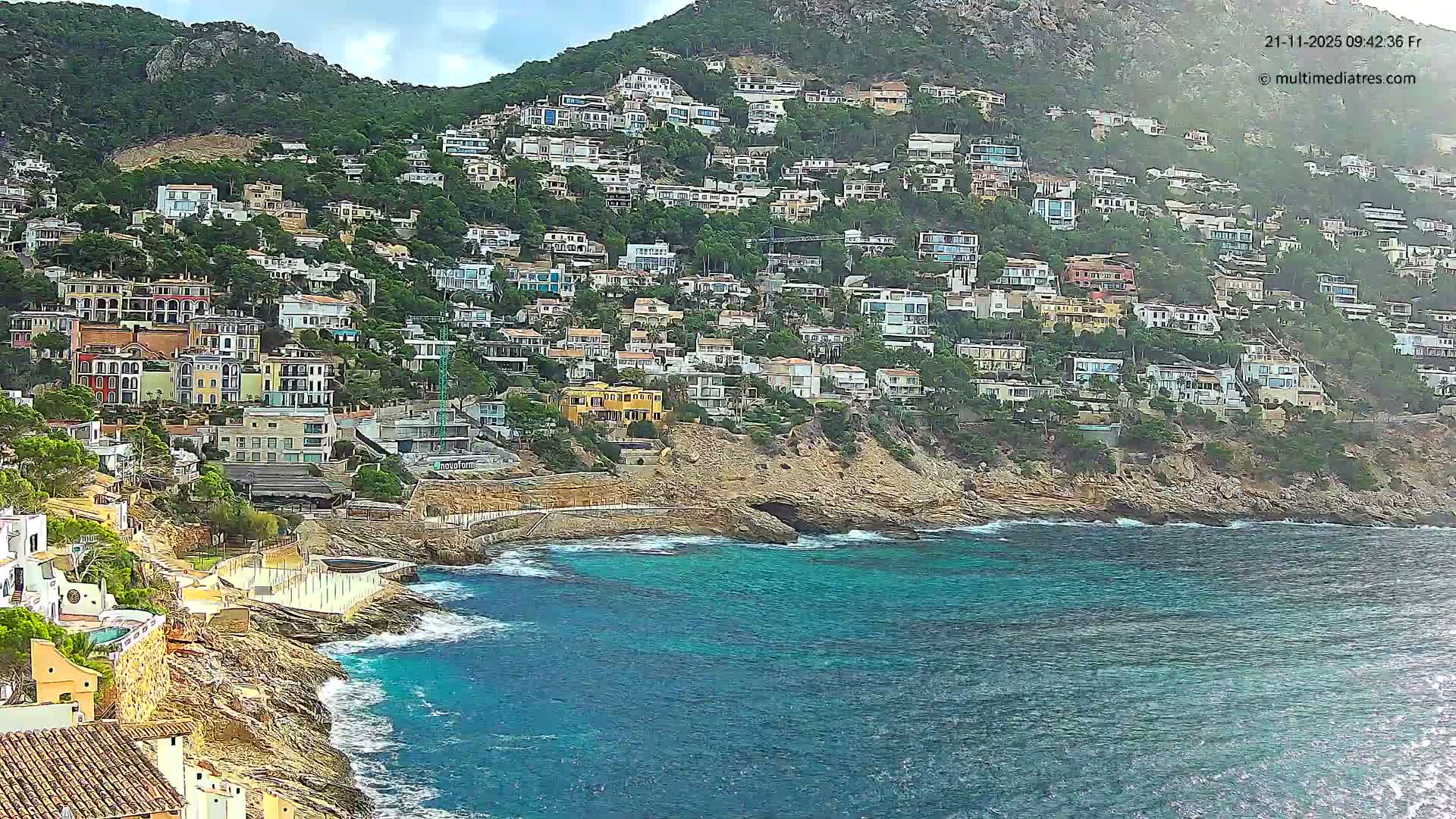 A steep, green hillside densely packed with colorful houses descends to a rocky coastline where clear blue-green waters meet the shore under a partly cloudy sky.