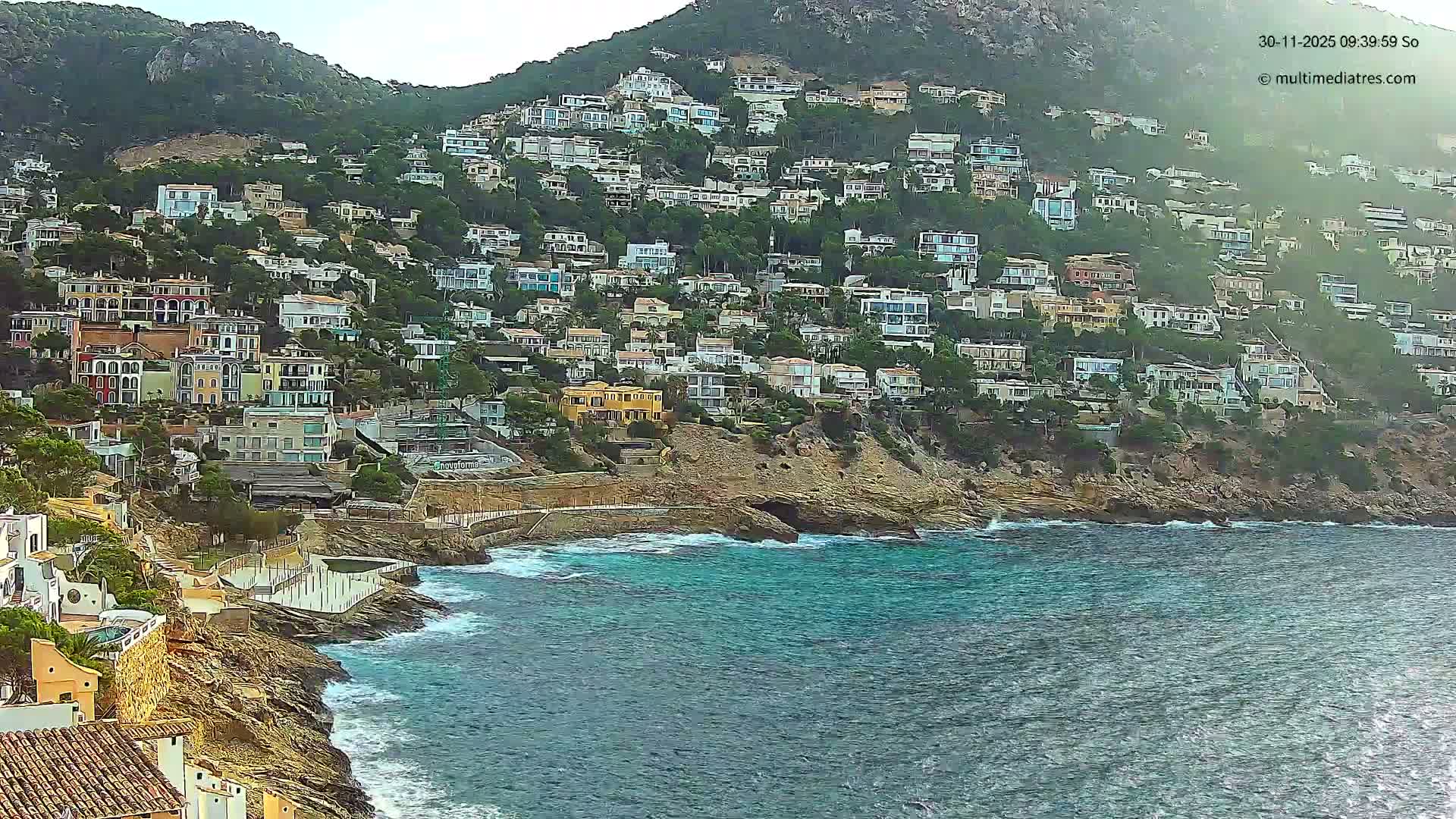 A vibrant coastal town with numerous houses sprawling up a steep, tree-covered mountain overlooks a turquoise sea with gentle waves under a bright, clear sky.