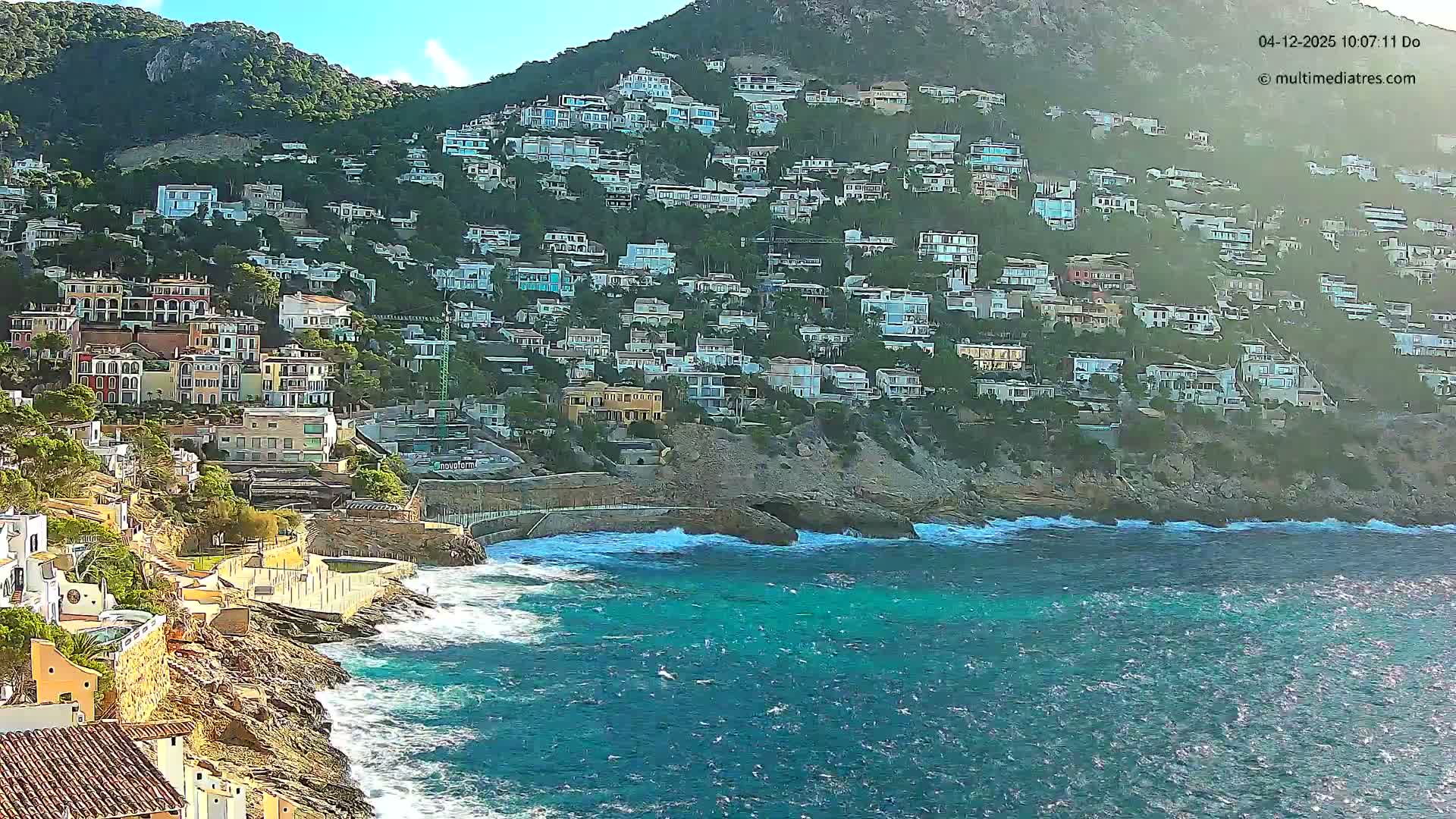 Numerous white and colorful houses dot a steep, lush green mountainside that plunges into a vibrant blue-green sea with waves crashing against a rocky coast and a small concrete beach, all under a clear, sunny sky.