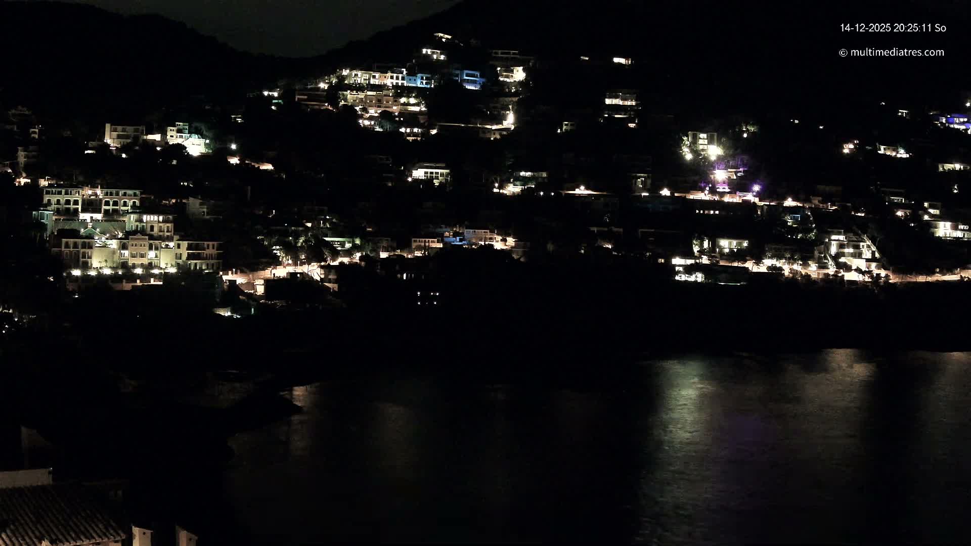 Numerous white and colorful houses dot a steep, lush green mountainside that plunges into a vibrant blue-green sea with waves crashing against a rocky coast and a small concrete beach, all under a clear, sunny sky.