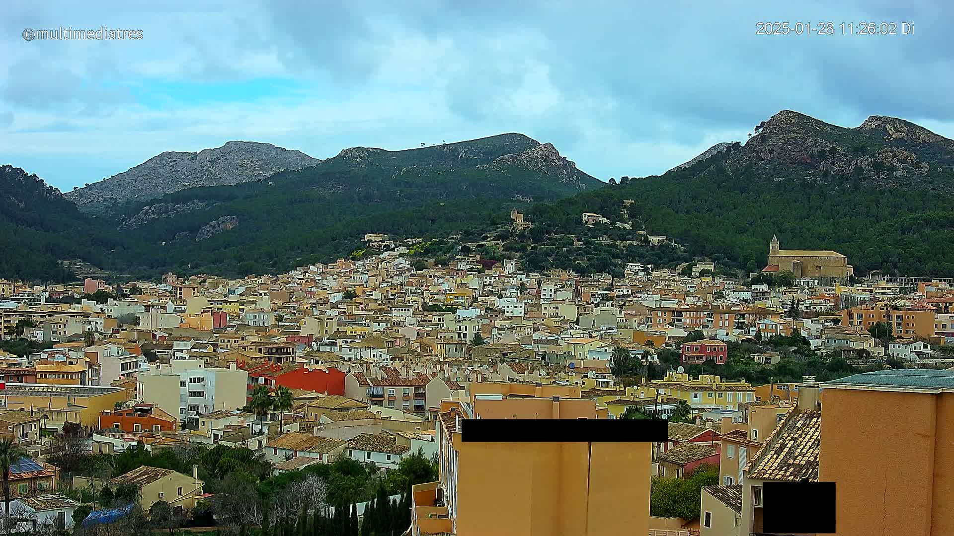 A sprawling town of closely packed buildings nestles in a valley surrounded by verdant, mountainous terrain under a mostly cloudy sky.