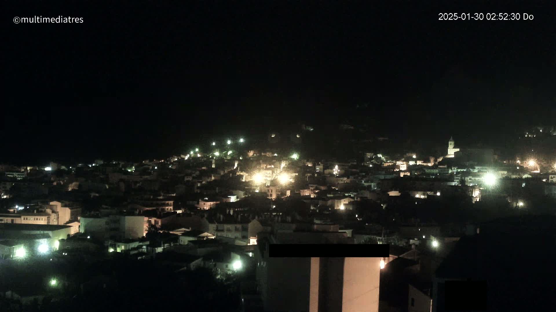 A nighttime, aerial view of a town illuminated by scattered lights, nestled on a hillside under a dark sky.