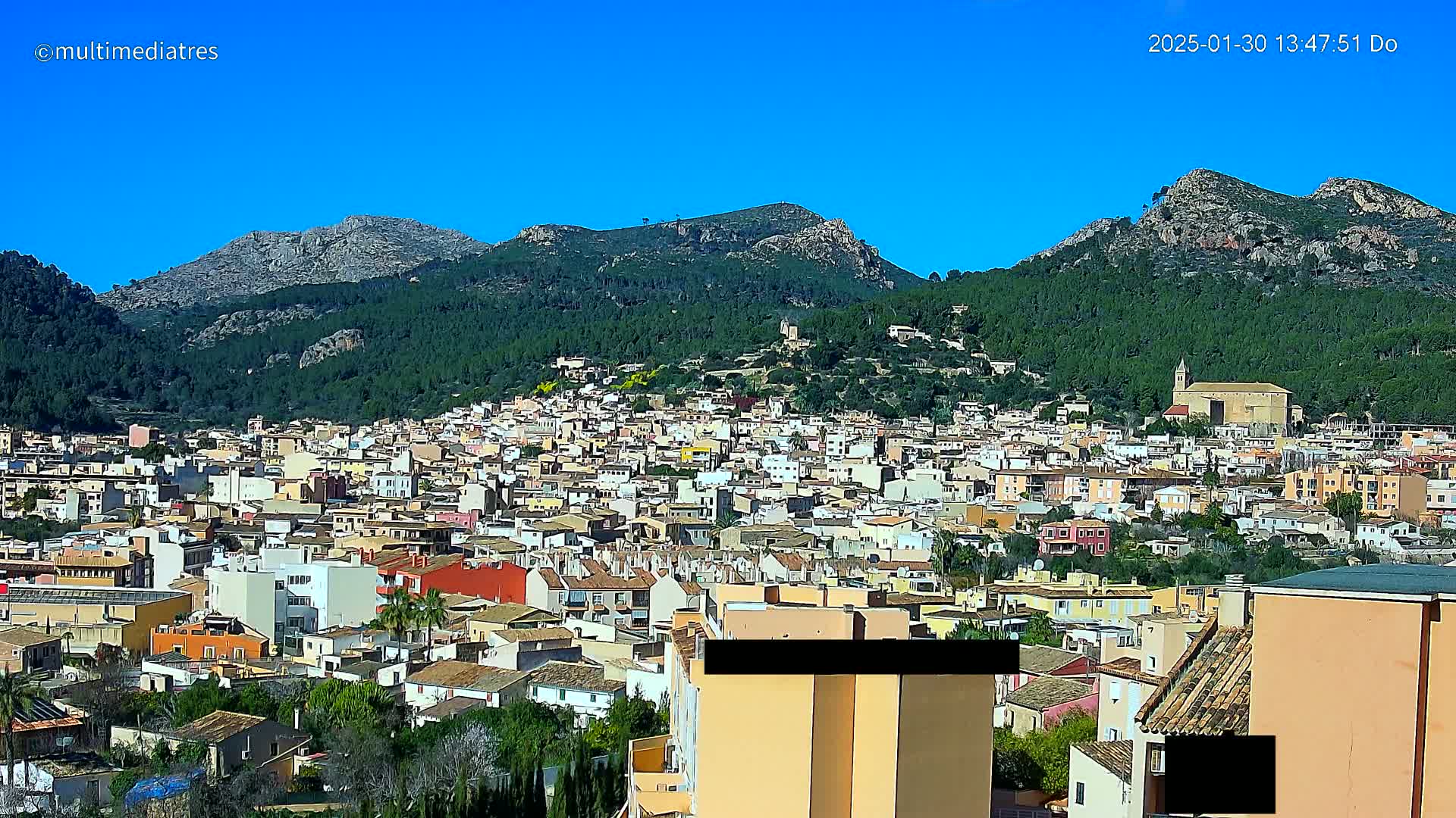 A sprawling town nestled in a valley, surrounded by verdant hills and mountains under a clear blue sky.