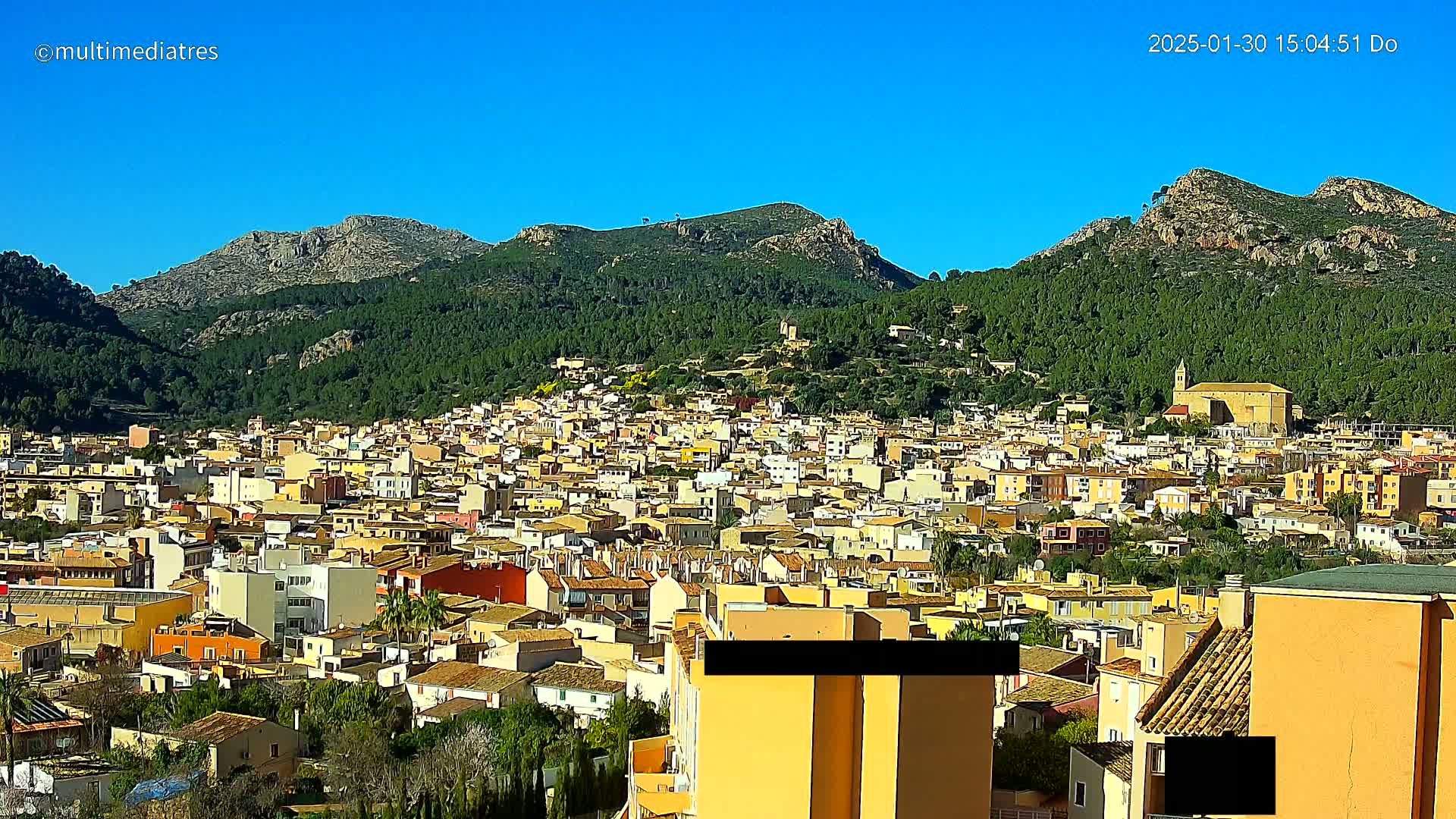 A sprawling town of tan and yellow buildings nestles at the base of a verdant mountain range under a clear blue sky.