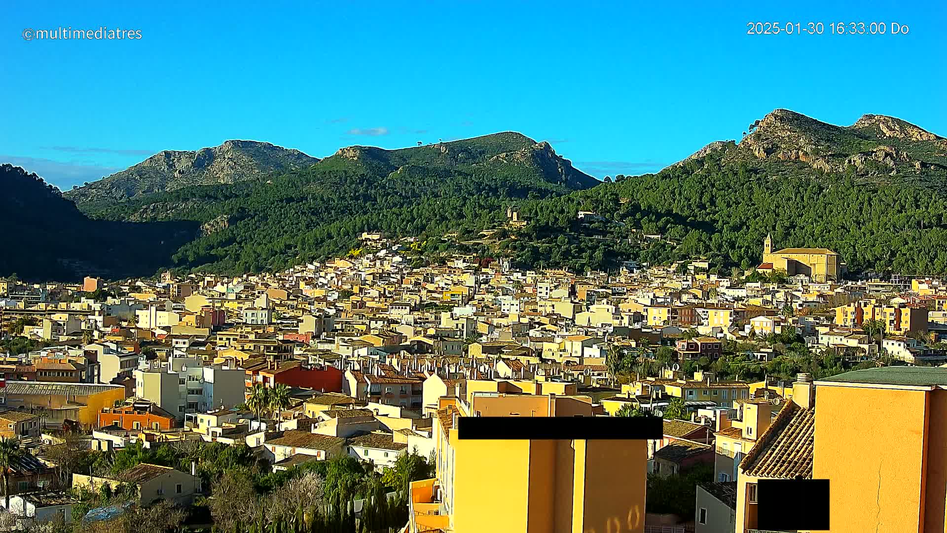 A sprawling town of closely packed, light-colored buildings nestles against a backdrop of verdant, mountainous terrain under a clear, bright blue sky.