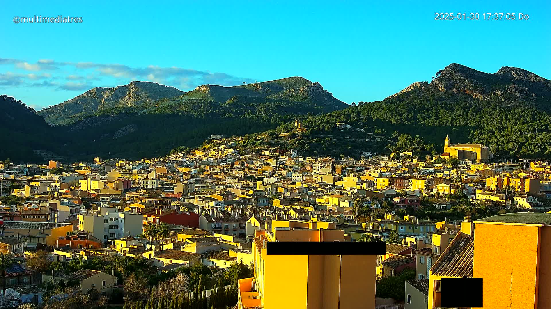 A sprawling town of yellow buildings nestled against a backdrop of verdant, sunlit mountains under a clear blue sky.