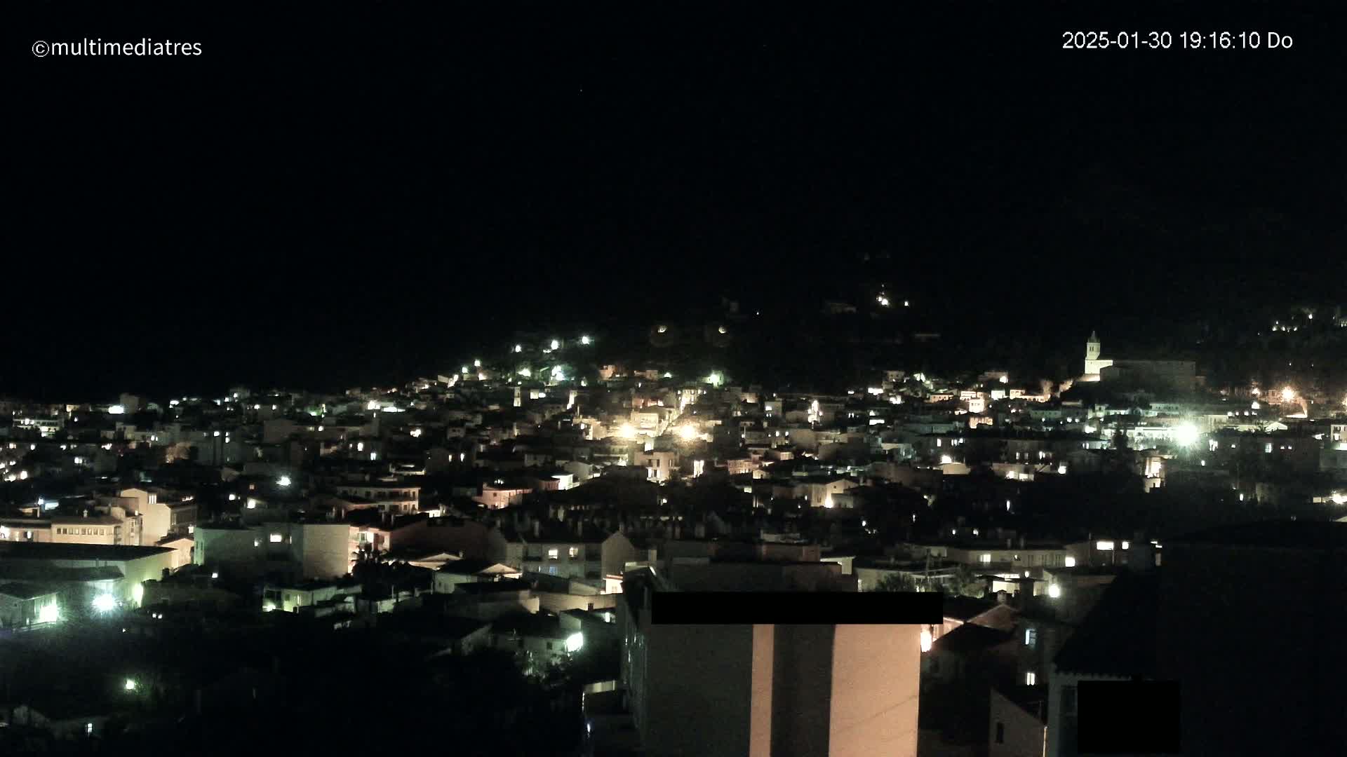 A nighttime, aerial view shows a town nestled on a hillside, illuminated by the lights of numerous buildings.